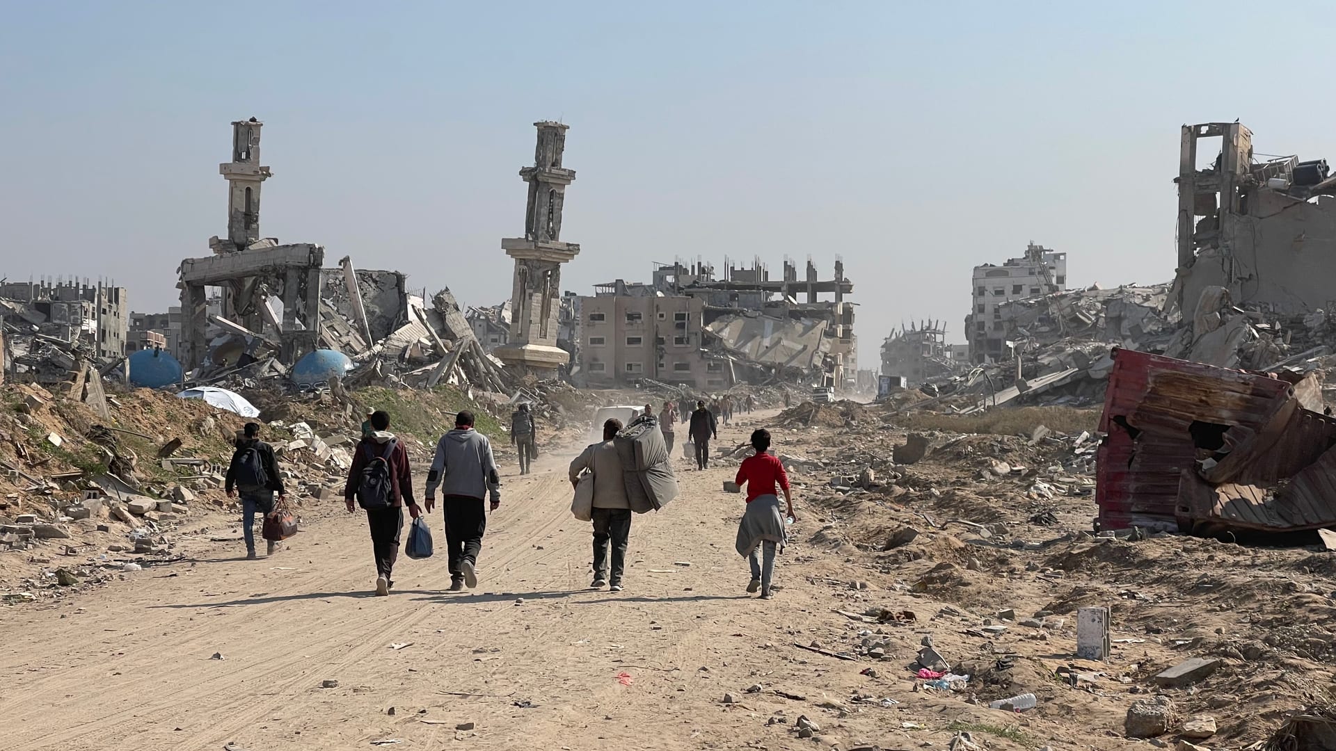 Un grupo de personas camina con bultos por un camino de tierra entre edificios y minaretes en ruinas en la Franja de Gaza.