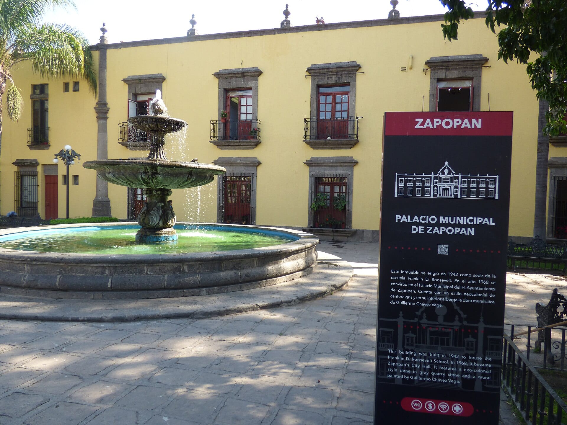 Fuente de piedra frente al edificio amarillo del Palacio Municipal de Zapopan, con un letrero informativo a la derecha.
