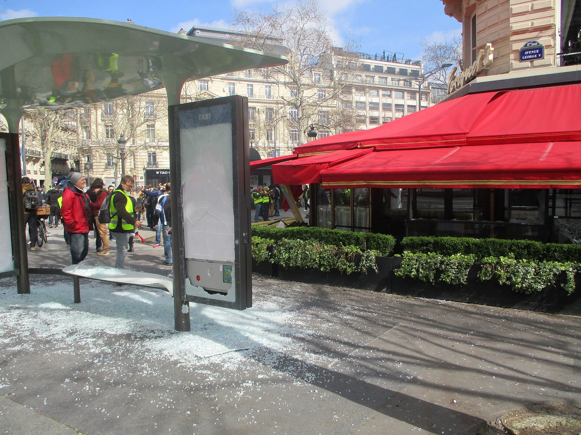 People in yellow vests walk past a bus shelter with shattered glass on the sidewalk, in front of a cafe with a red awning on a Paris avenue.