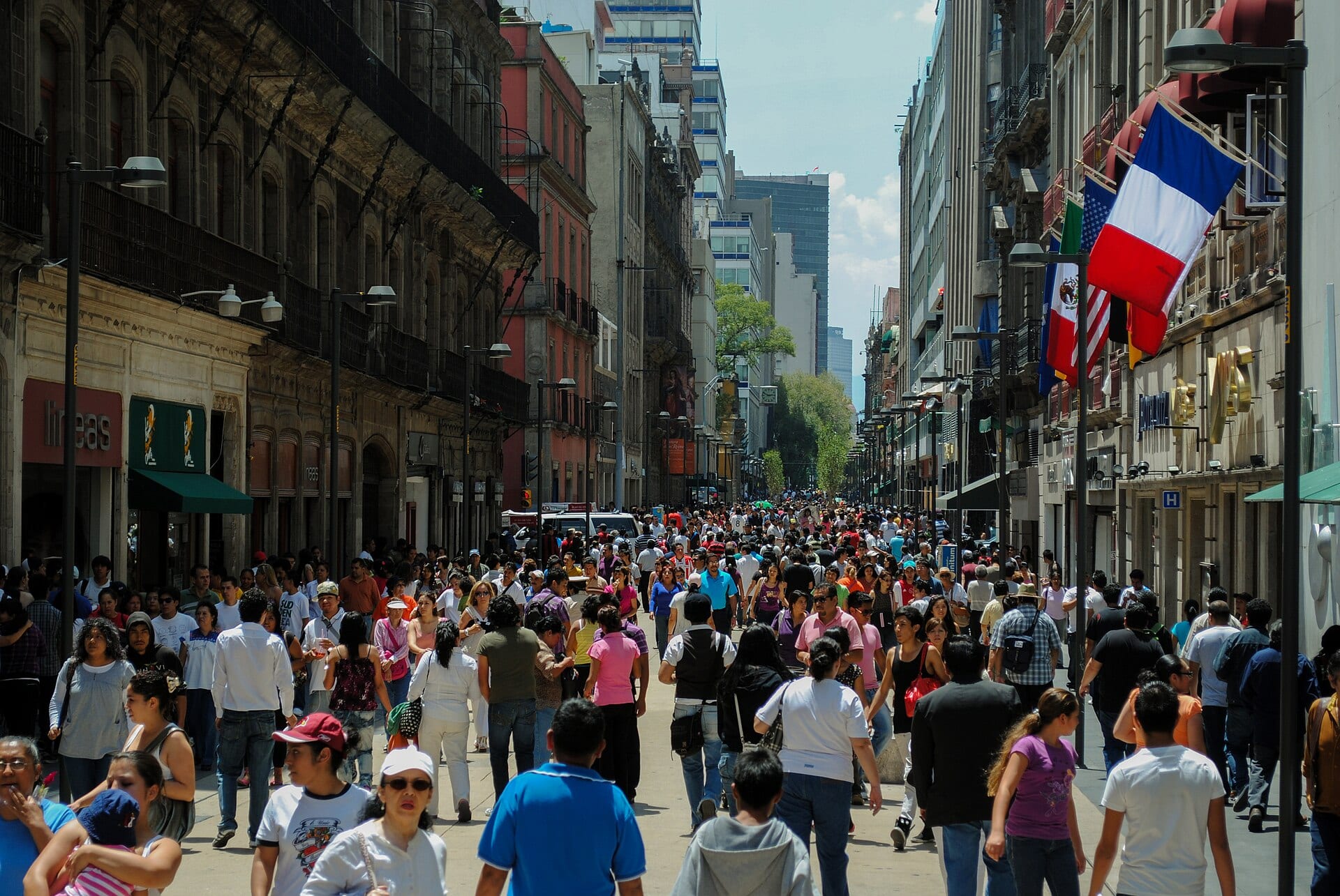 Una calle peatonal llena de gente entre edificios antiguos y modernos, con banderas colgadas a la derecha.