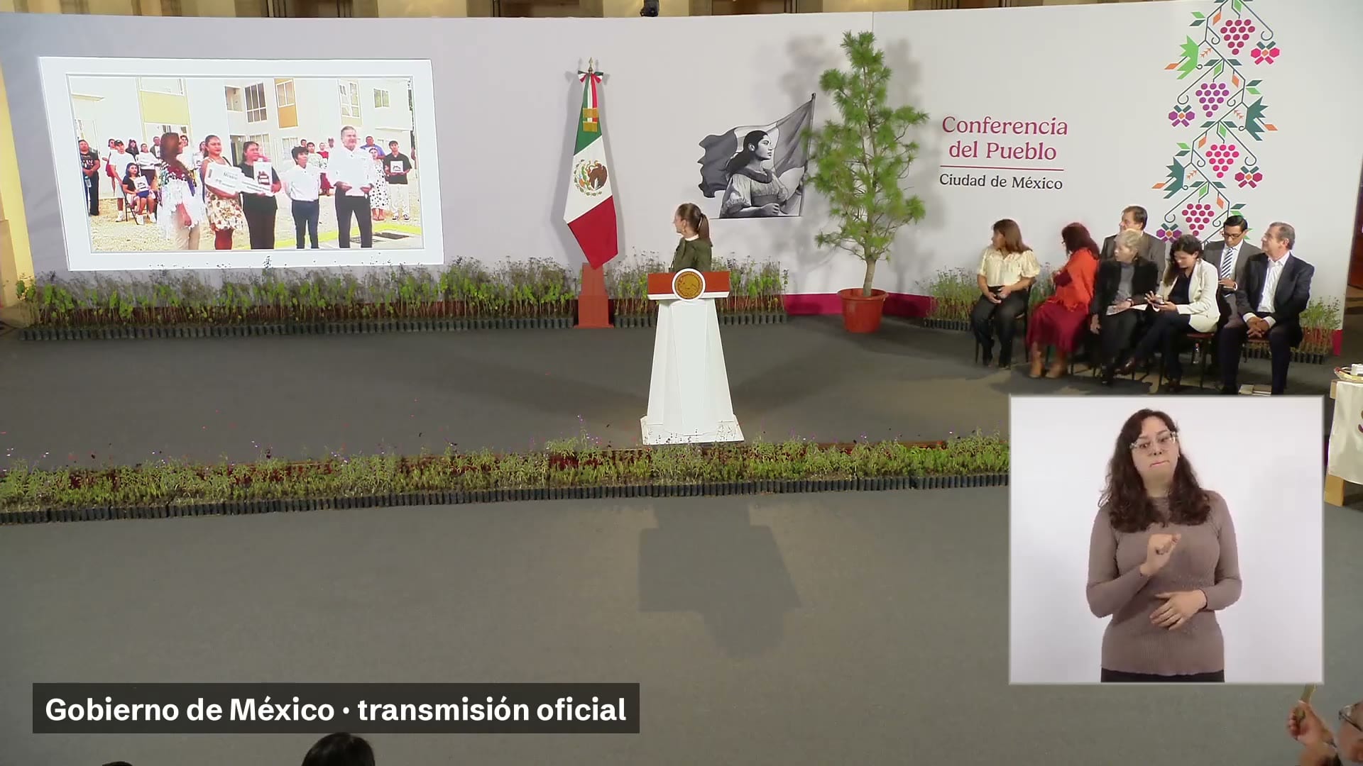 La presidenta Claudia Sheinbaum frente al podio de la conferencia, con una pantalla que muestra a familias frente a un conjunto de viviendas y, a la derecha, personas sentadas.