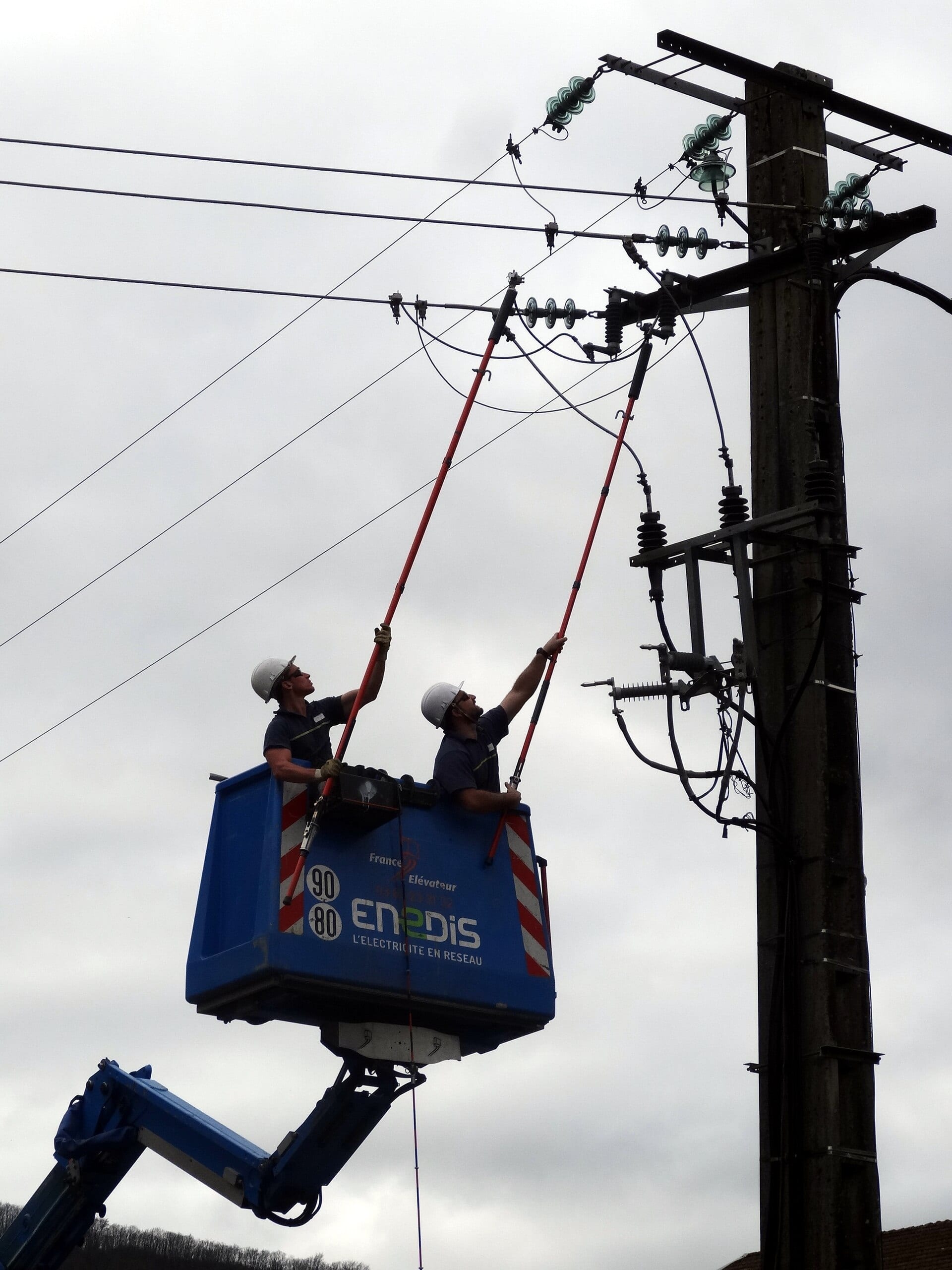 Two workers in hard hats use insulated poles from a blue Enedis lift basket beside a utility pole carrying power lines.