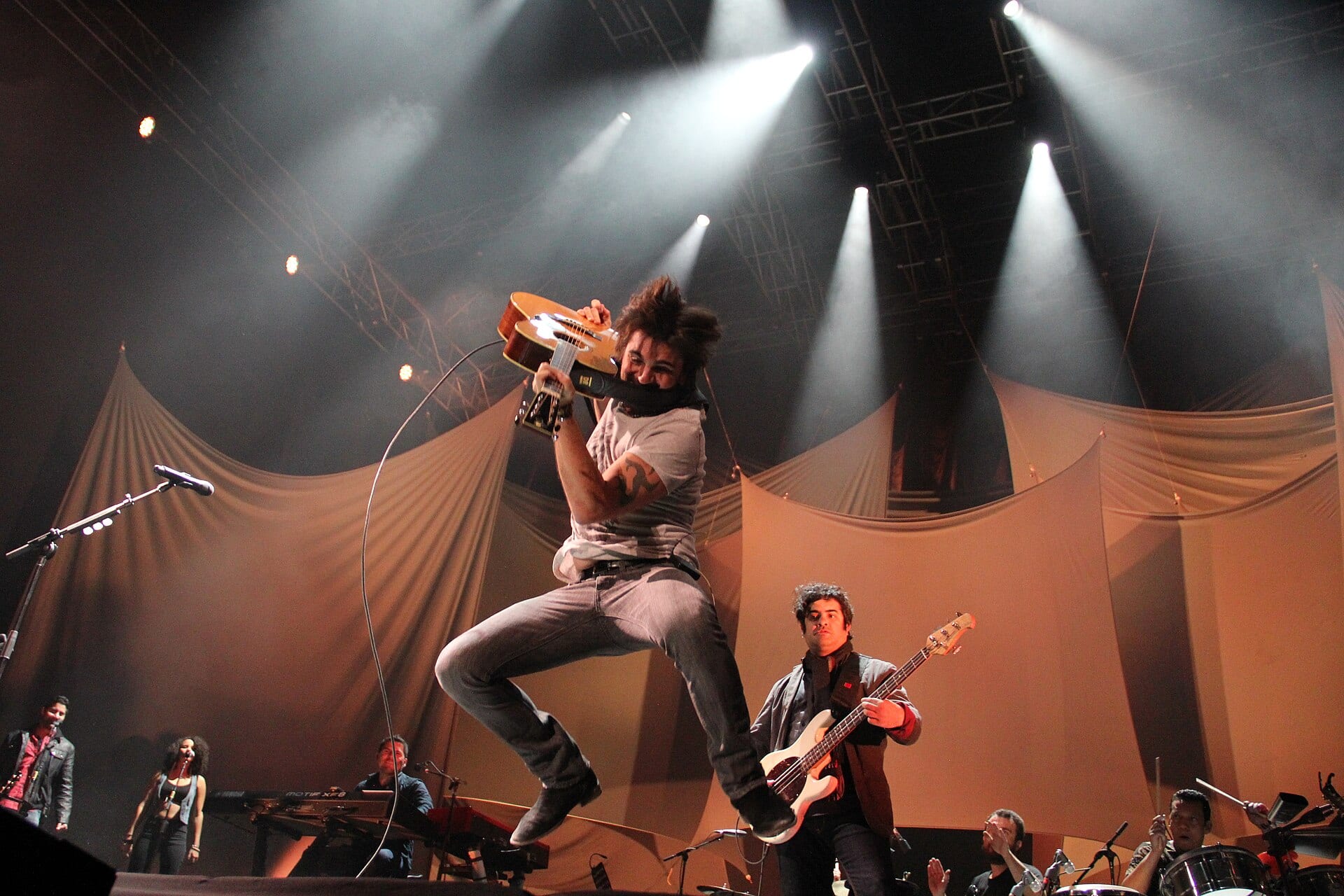 Juanes salta con la guitarra acústica en alto sobre un escenario iluminado por reflectores, con su banda detrás.