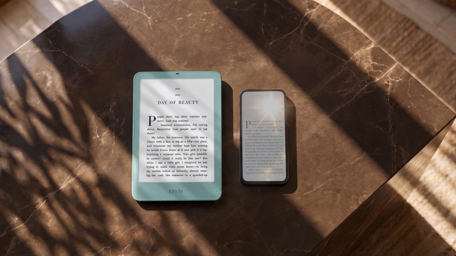 Un Kindle verde azulado con texto en pantalla junto a un teléfono con el sol reflejado en la pantalla, sobre una mesa de mármol oscuro con sombras de hojas.