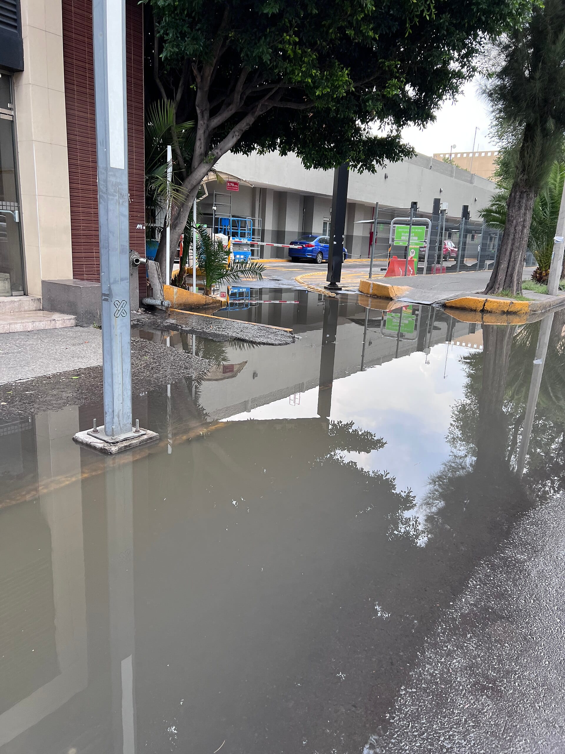 Un charco cubre parte de la calle y de la banqueta frente a un edificio, con árboles y un estacionamiento al fondo.