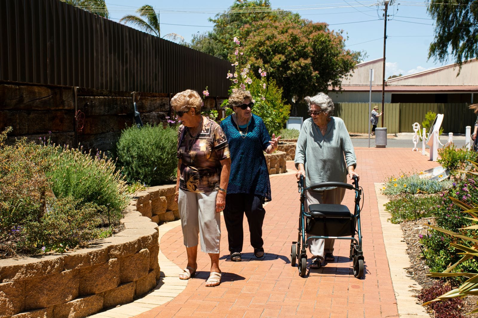 Tres mujeres mayores caminan por un sendero de ladrillo entre jardineras, una de ellas apoyada en un andador con ruedas.