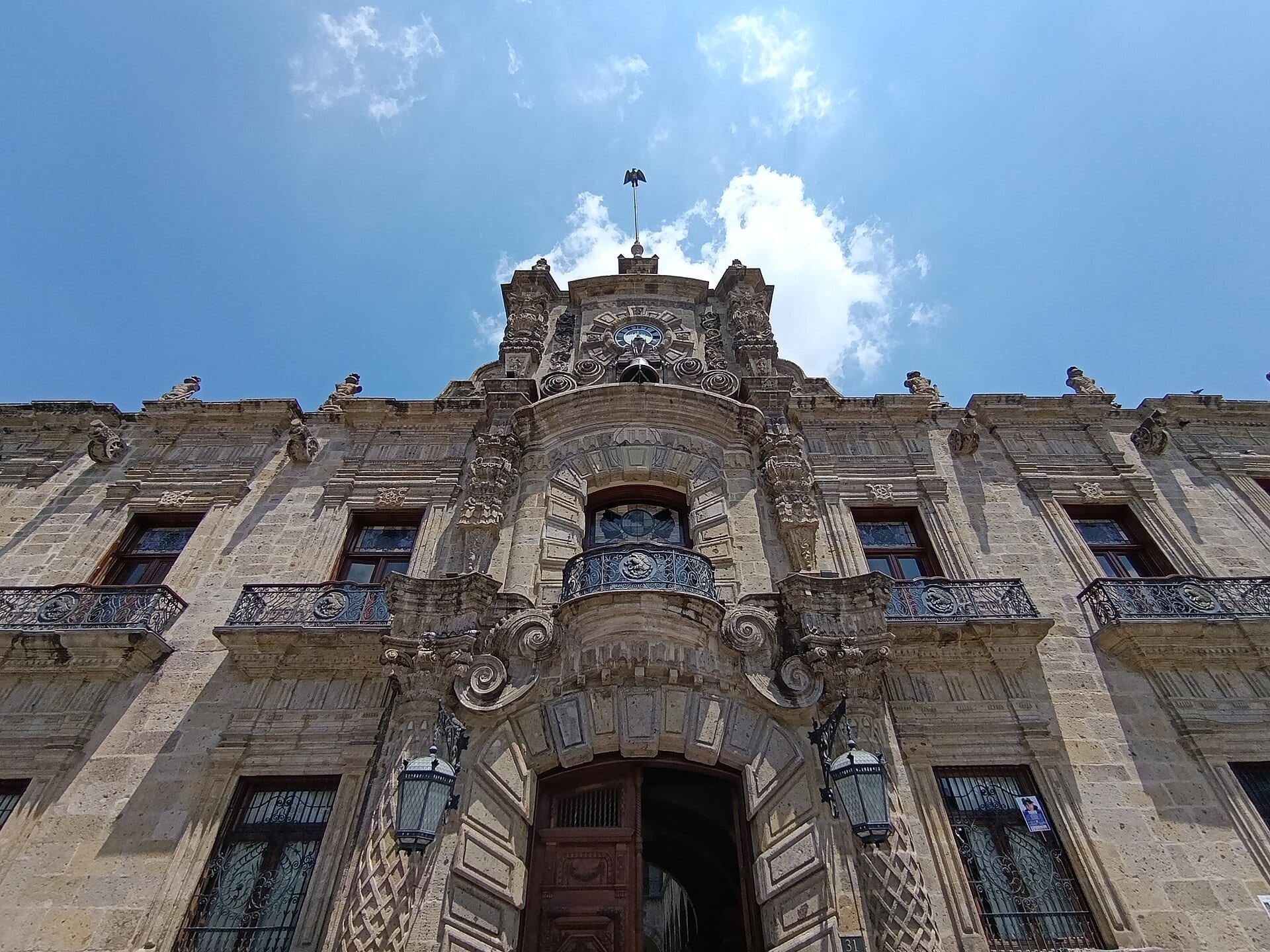 Fachada de cantera del Palacio de Gobierno de Jalisco vista desde abajo, con su balcón central, el reloj y un cielo azul con nubes.