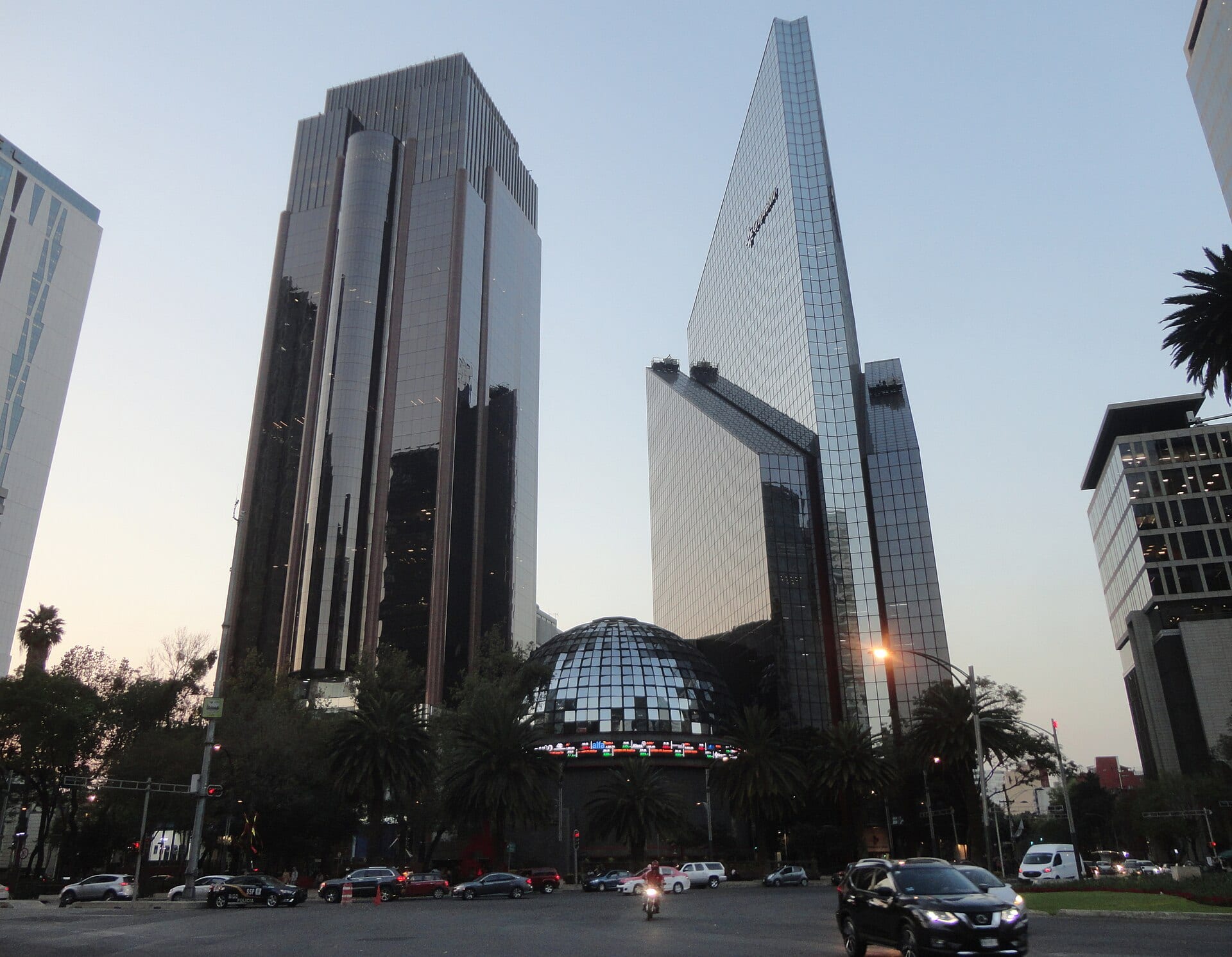 El domo de vidrio de la Bolsa Mexicana de Valores con su pantalla de cotizaciones, entre dos torres de cristal sobre Paseo de la Reforma al atardecer.