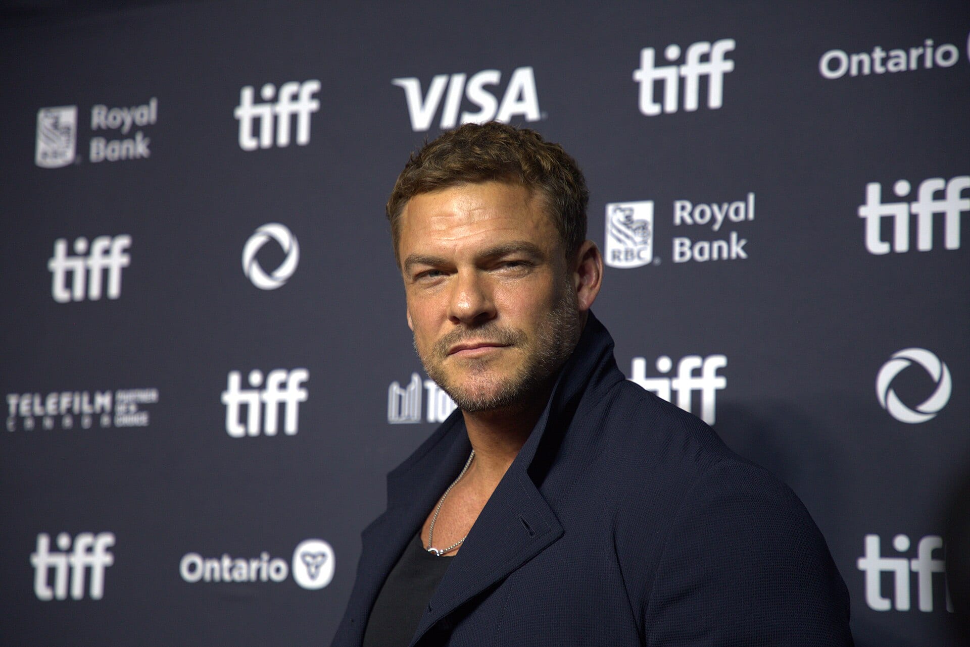 Alan Ritchson, con saco azul marino y camiseta negra, posa de perfil ante un fondo con logotipos del festival TIFF.