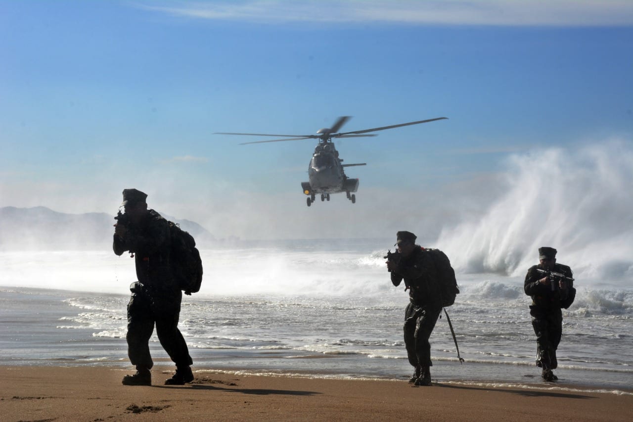 Three troops with rifles advance along a beach as a helicopter approaches over the surf.