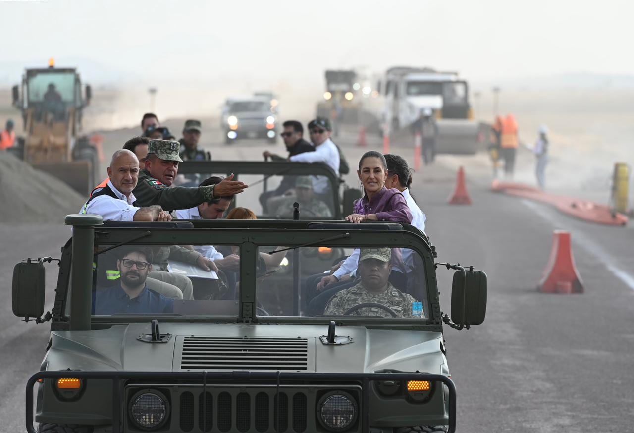 La presidenta Sheinbaum y funcionarios recorren en un vehículo militar abierto una obra con maquinaria y conos naranjas.