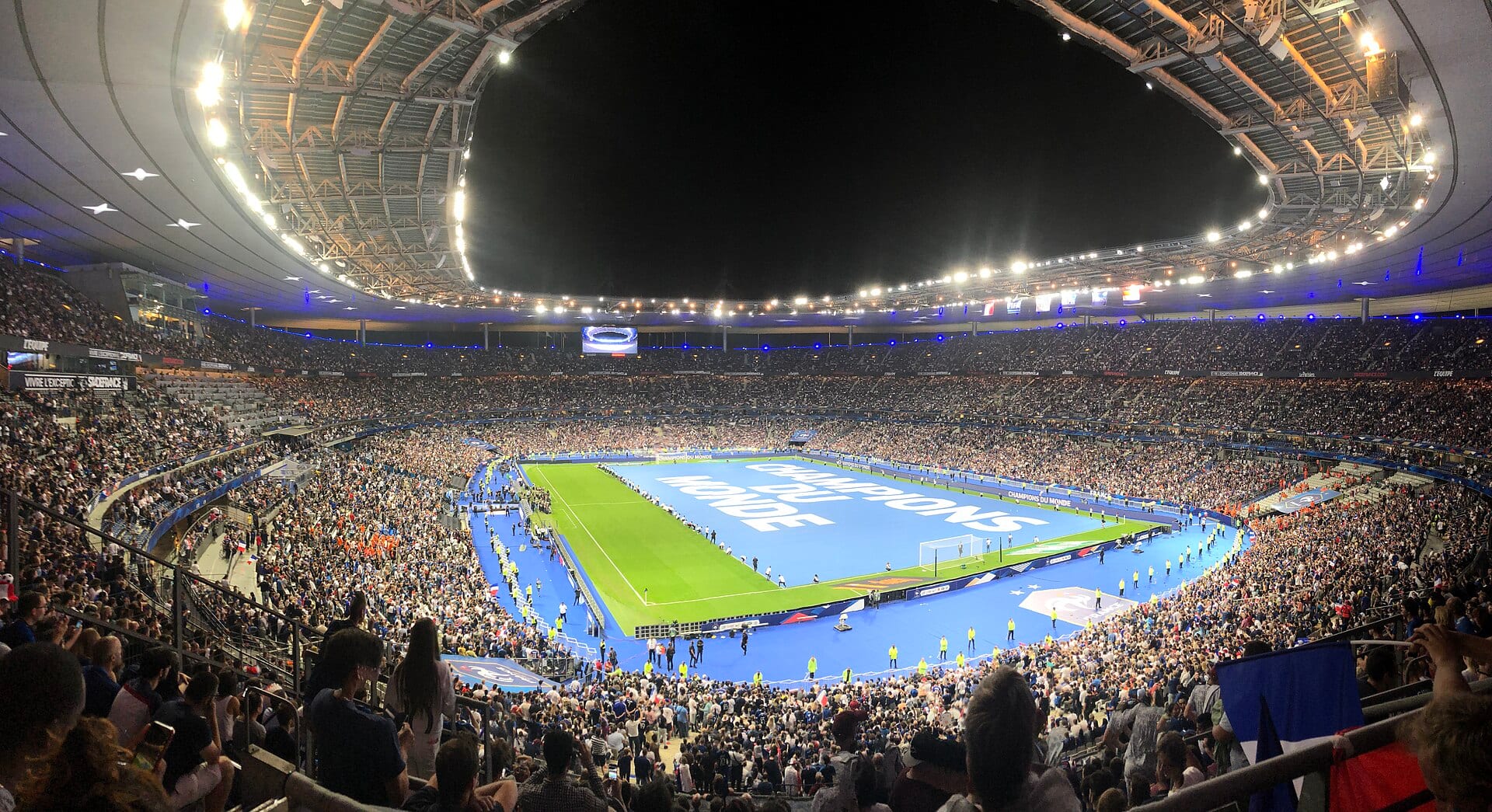 Panorámica nocturna del Stade de France lleno, con el césped cubierto por una lona azul con la leyenda Champions du Monde.