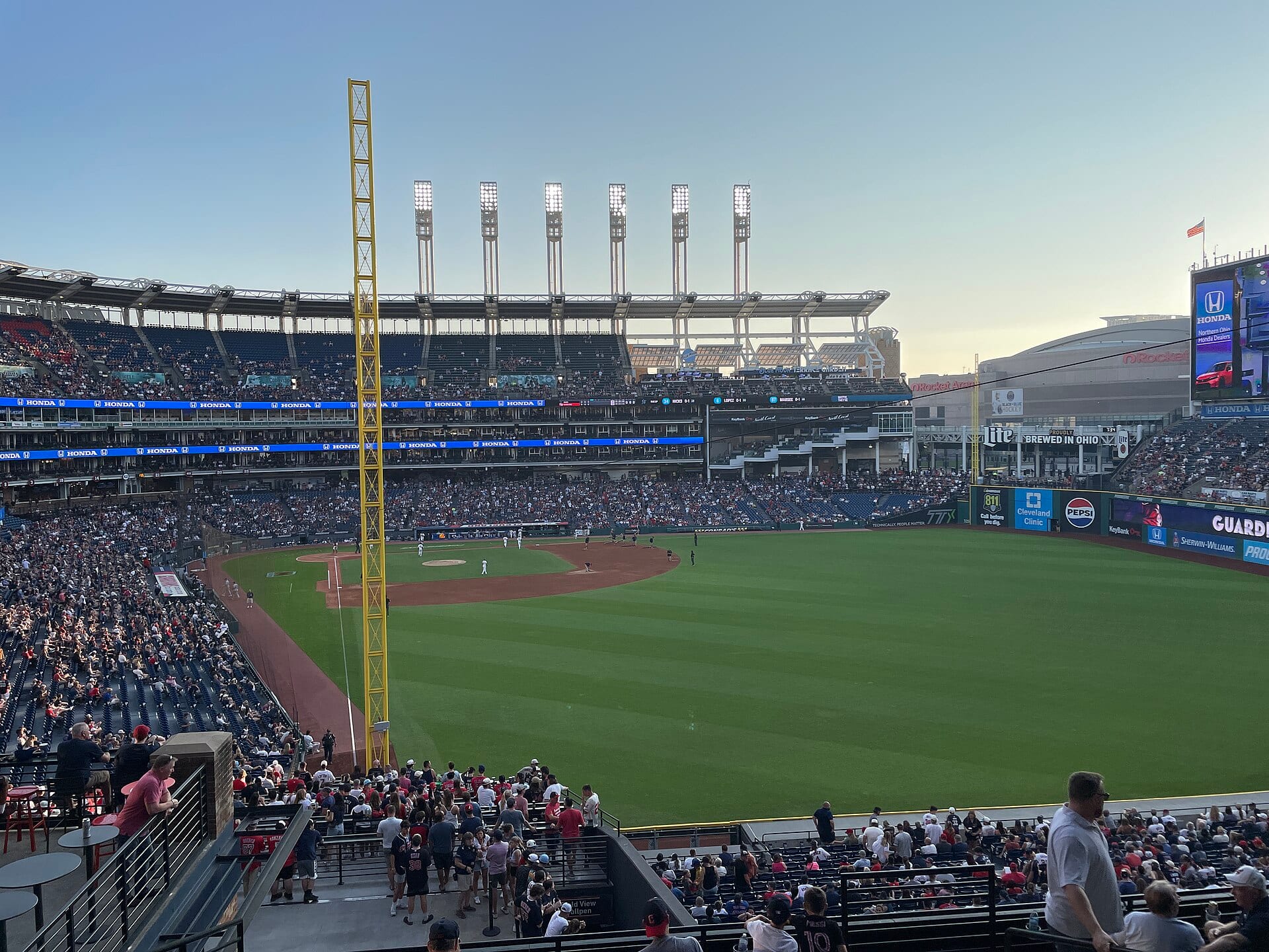 Vista del Progressive Field desde las gradas del jardín izquierdo, con el diamante al fondo, las torres de luces y público en los asientos al atardecer.