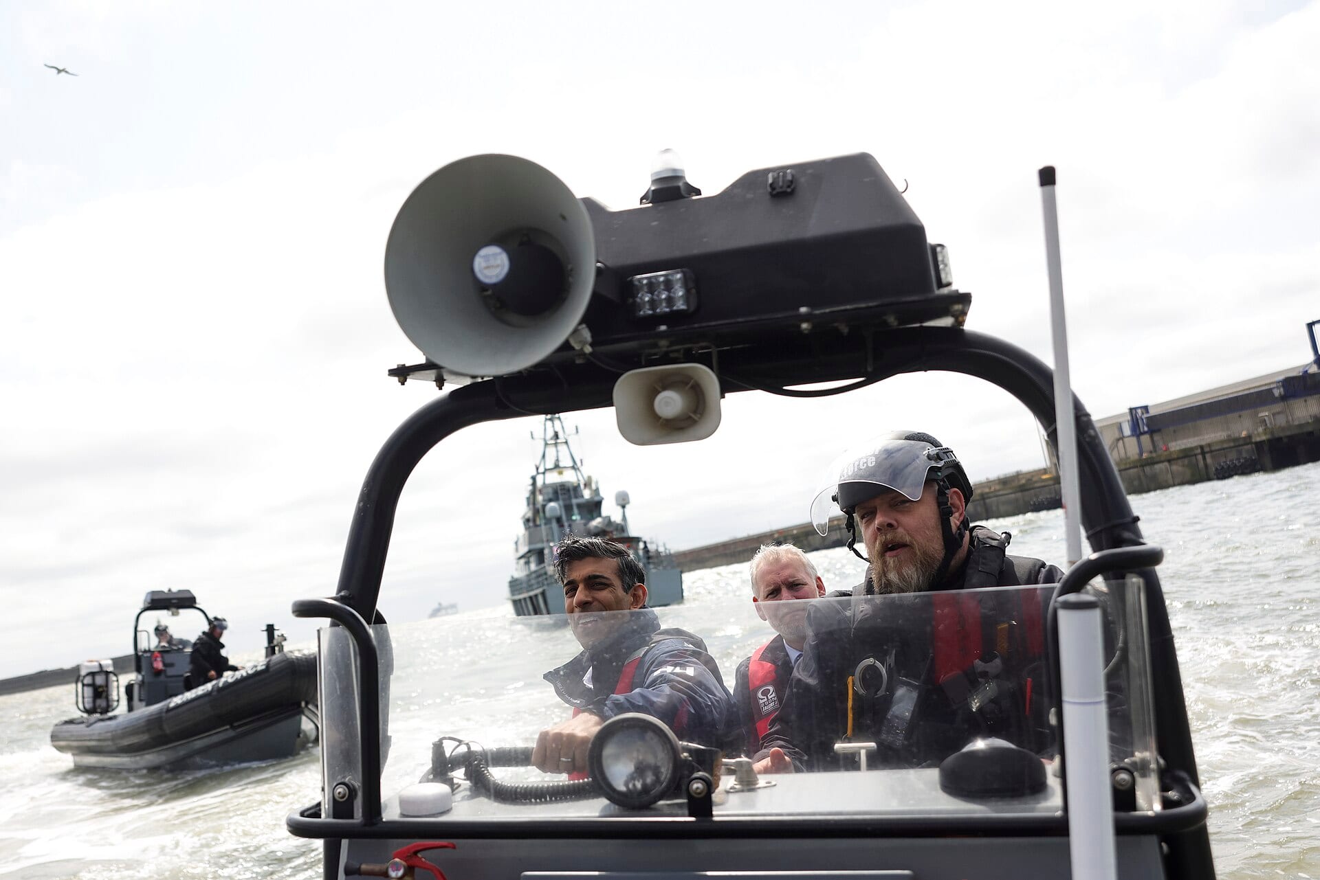 Tres hombres con chalecos salvavidas, dos de ellos con casco, viajan en una lancha neumática de la Fuerza Fronteriza con un altavoz al frente y un buque patrulla al fondo.