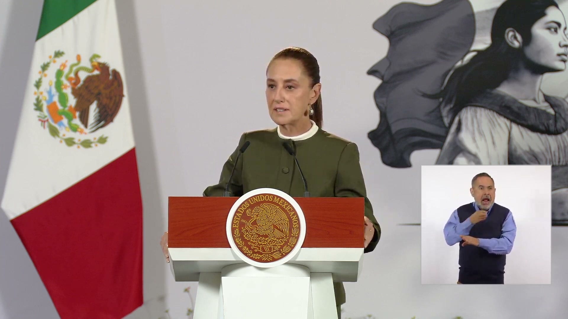 La presidenta Claudia Sheinbaum habla detrás del atril con el escudo nacional, junto a una bandera de México.