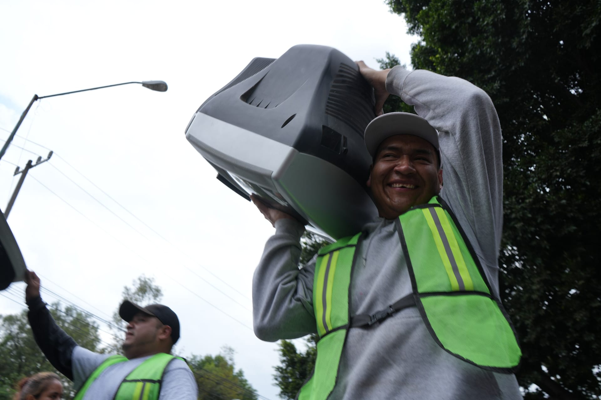 Un trabajador con chaleco verde carga un televisor antiguo sobre el hombro y sonríe.