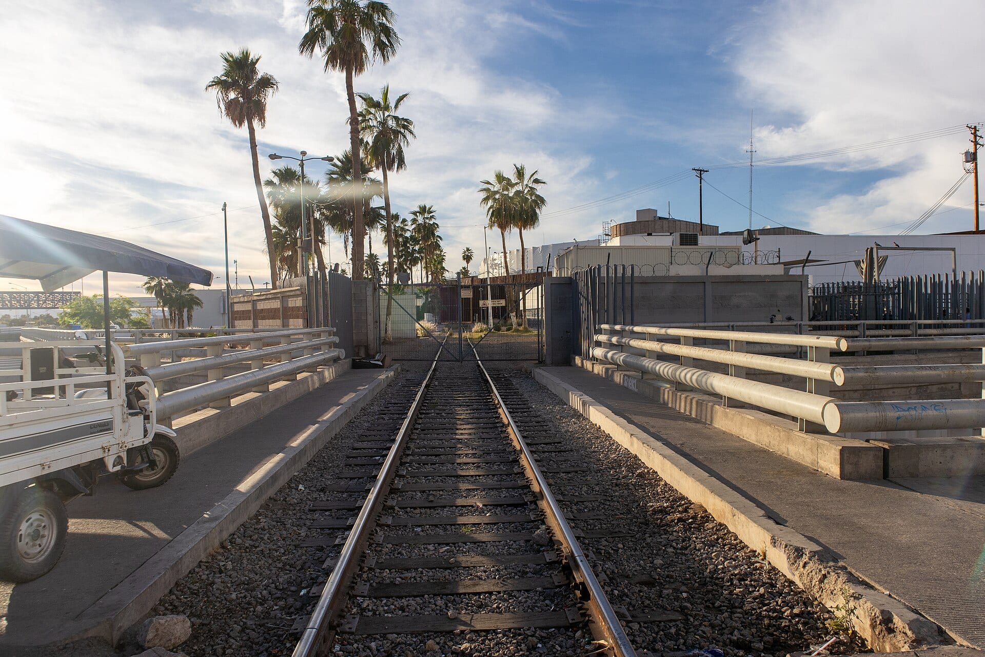Vías de tren entre dos pasillos con barandales que llegan a una reja, con palmeras y edificios de la aduana de Mexicali al fondo.