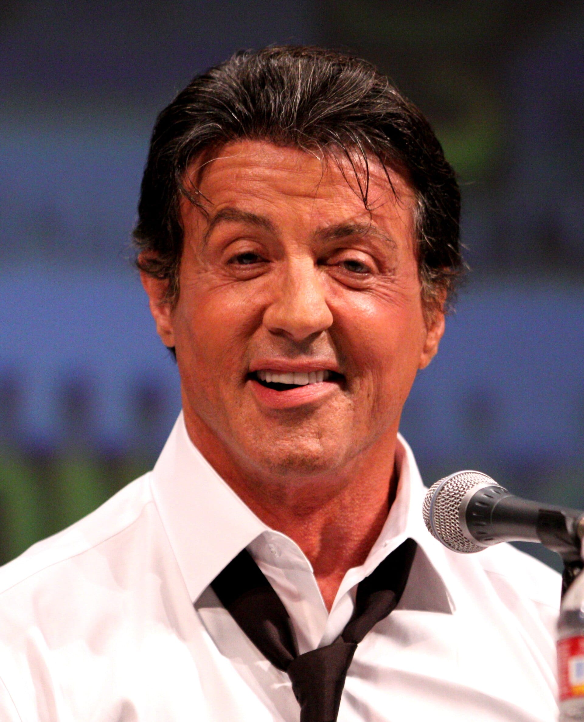 Sylvester Stallone sonríe mientras habla frente a un micrófono, con camisa blanca y corbata negra floja, sobre un fondo azul desenfocado.