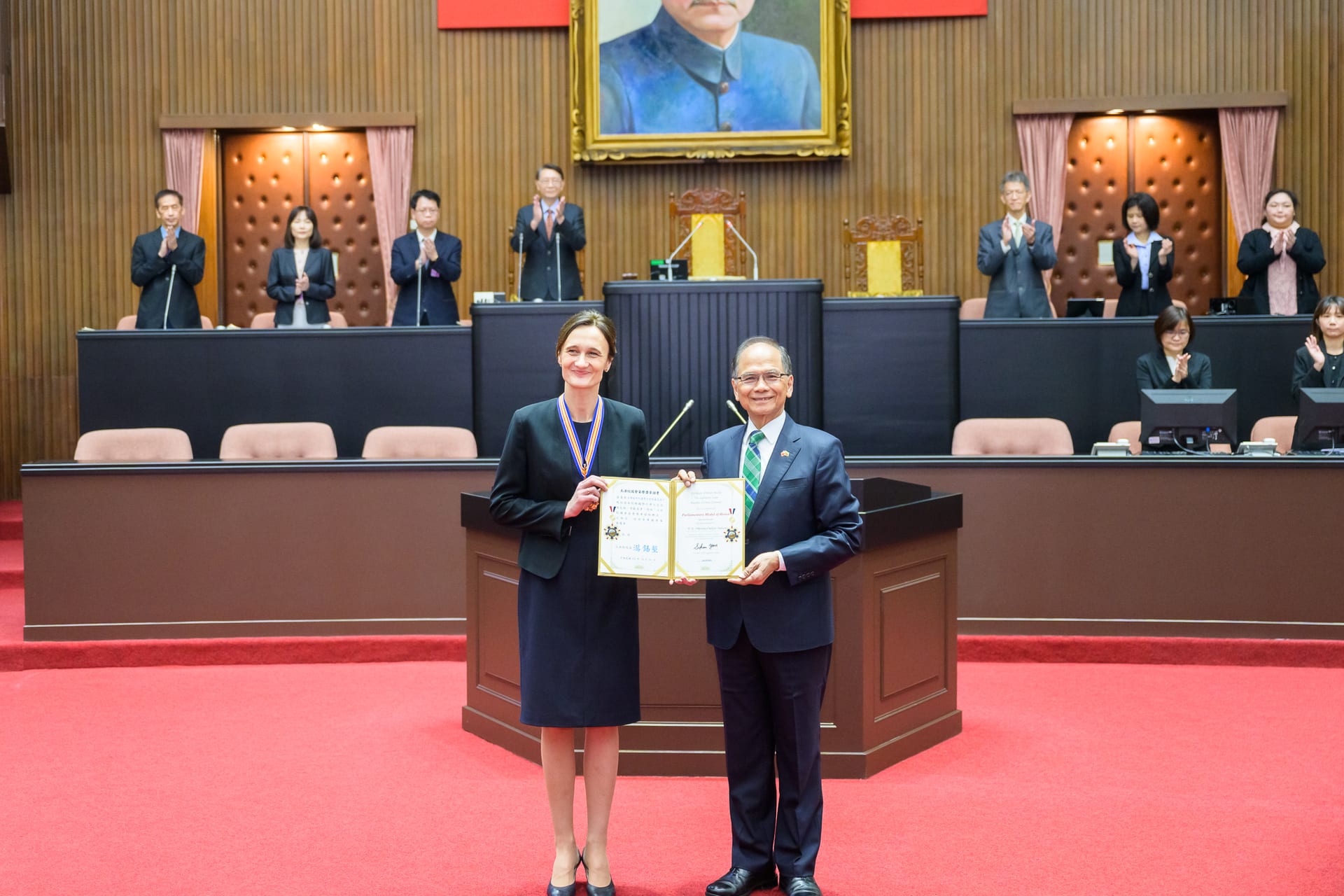 Una mujer y un hombre sostienen juntos un diploma enmarcado en el salón del pleno del Yuan Legislativo, con alfombra roja y personal aplaudiendo al fondo.
