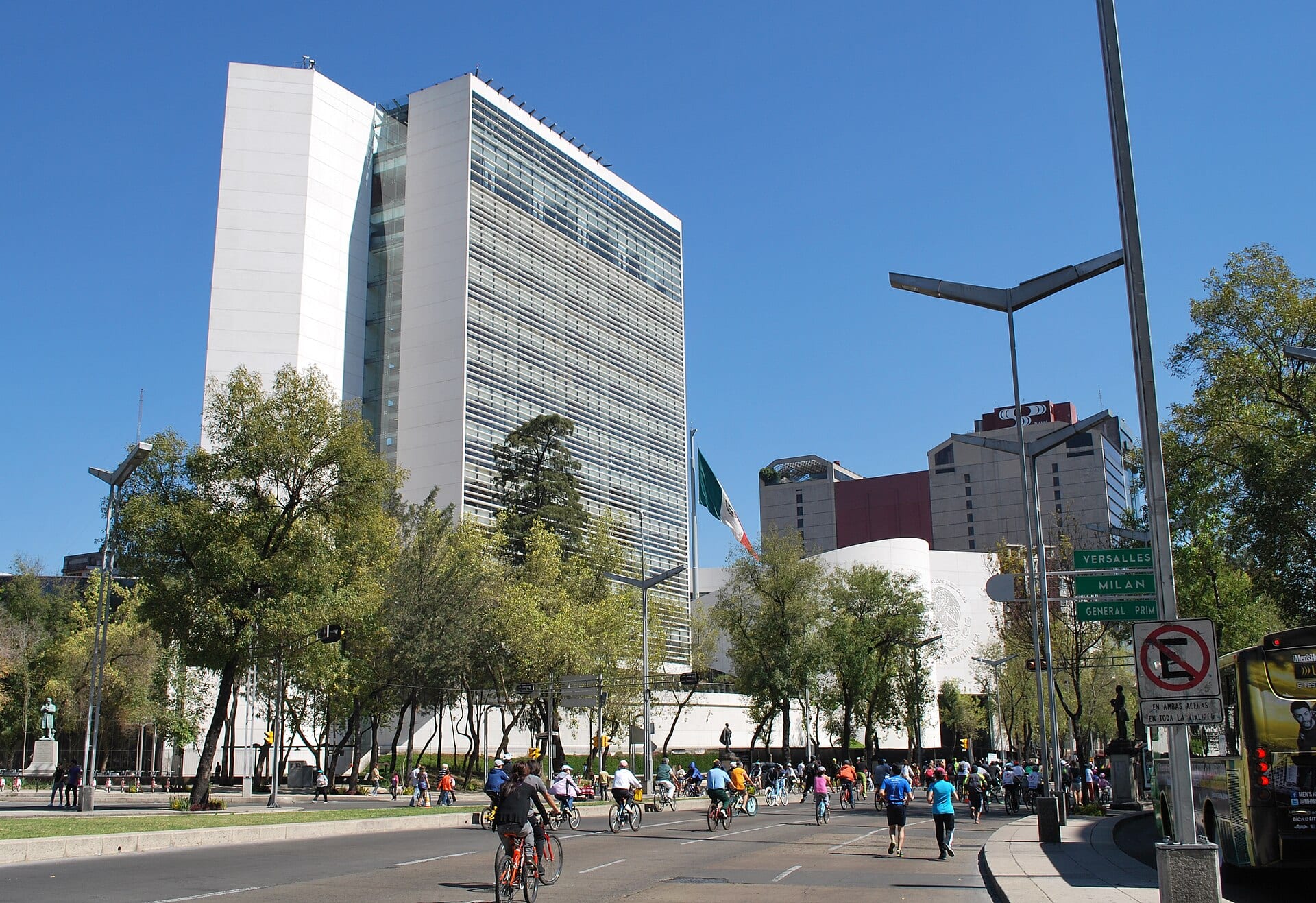 Torre blanca del Senado de la República en una avenida con árboles y ciclistas en primer plano, bajo un cielo azul.