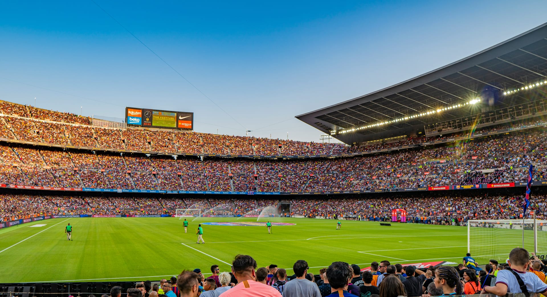 Estadio lleno de aficionados bajo un cielo despejado al atardecer; en el césped, cuidadores caminan entre aspersores y en primer plano hay público de pie.
