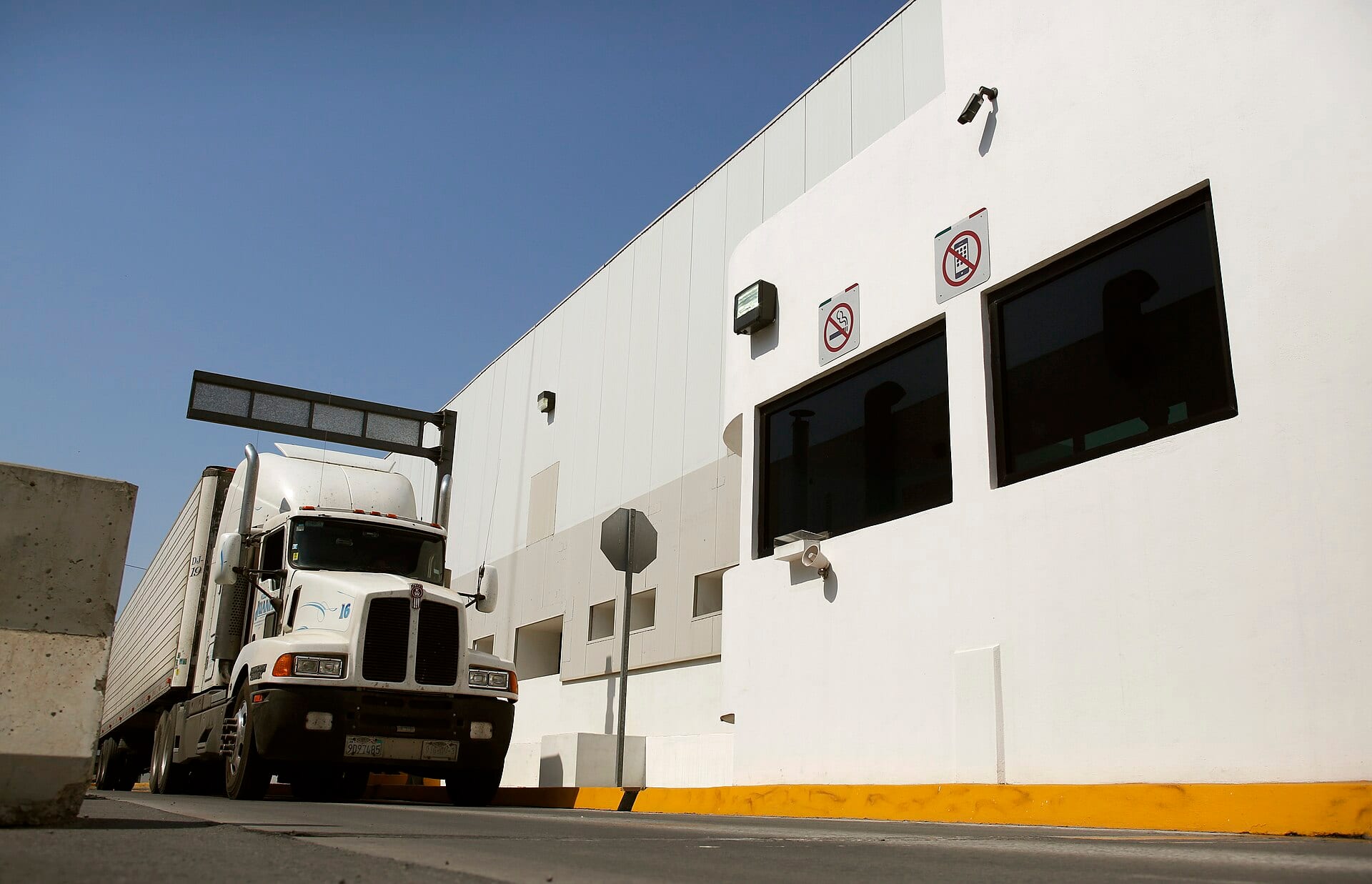A white tractor-trailer drives past a white inspection booth at a cargo border crossing.