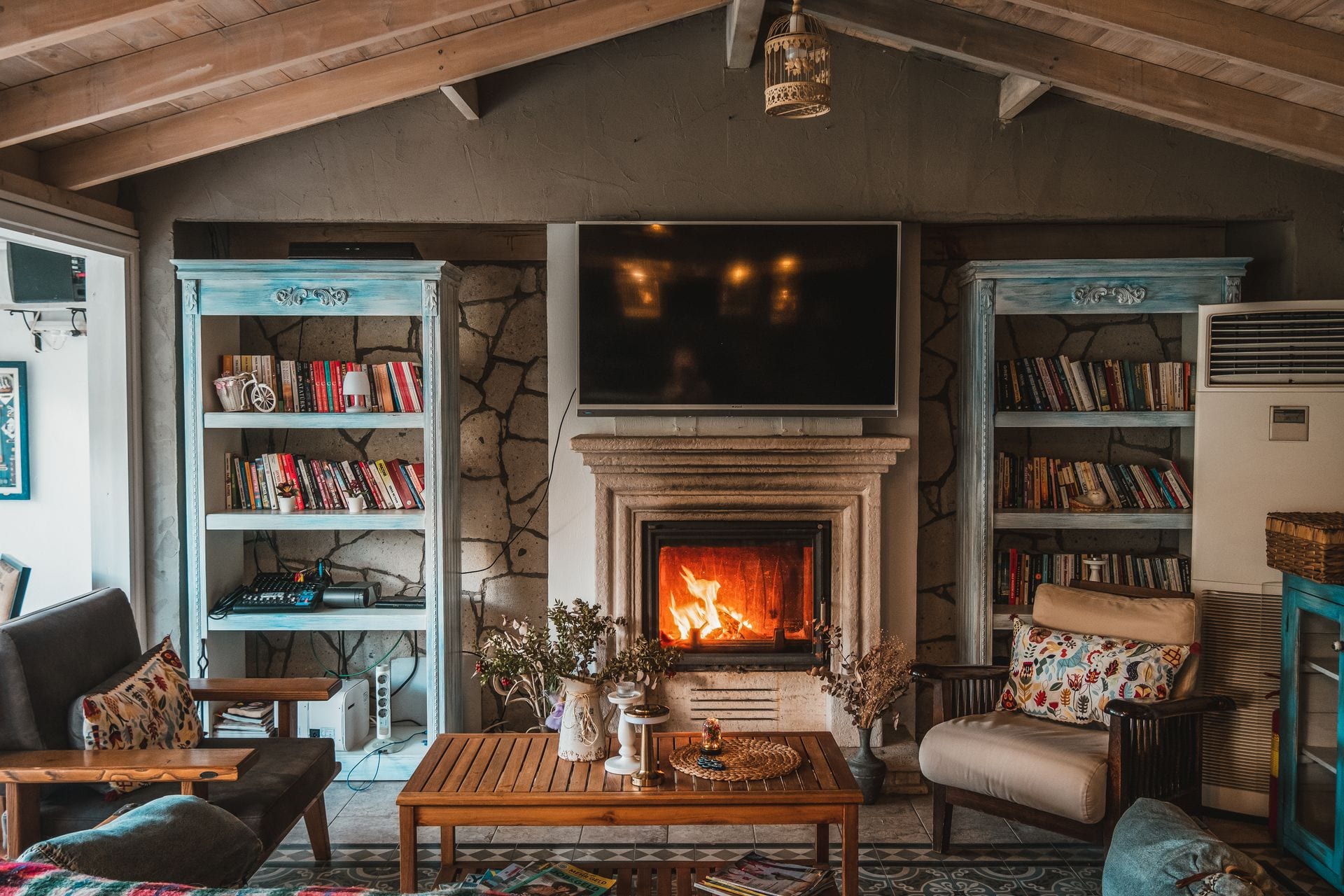 Sala con chimenea encendida, dos libreros azules a los costados, una mesa de madera al centro y sillones con cojines de colores.