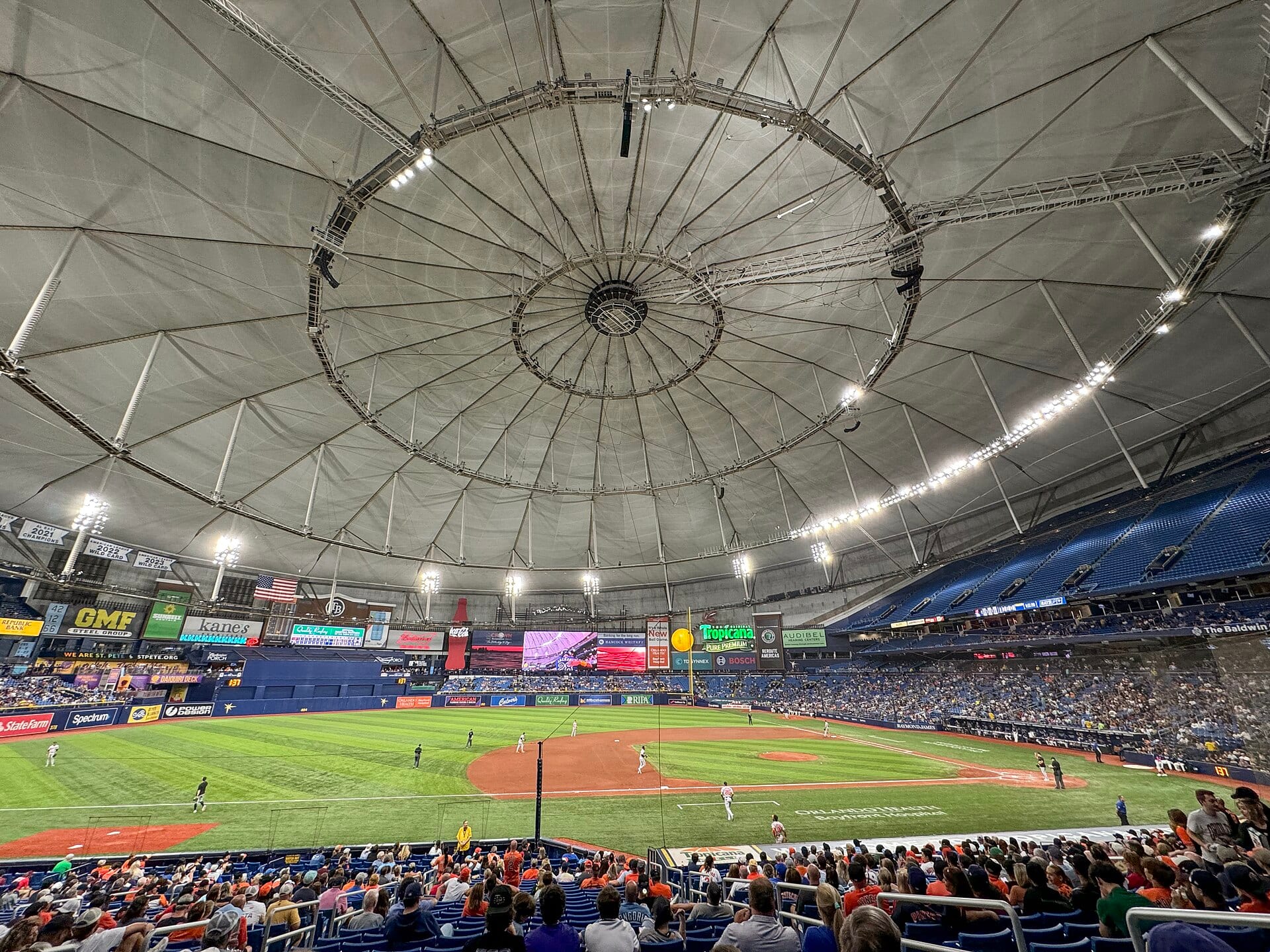 Interior del Tropicana Field desde la grada baja, con el techo de lona, el diamante con jugadores en posición y las tribunas azules llenas de aficionados.