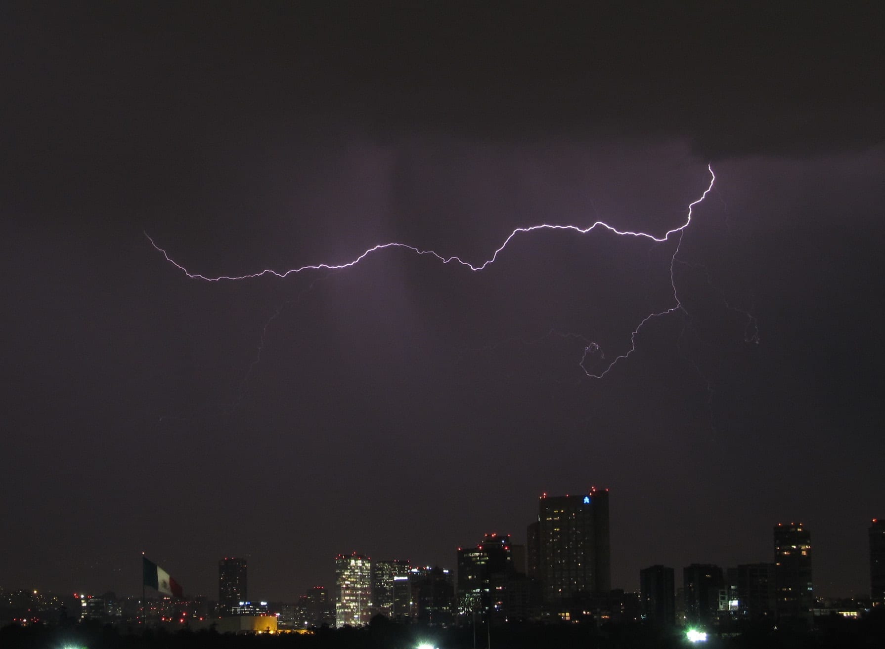 Un rayo cruza un cielo oscuro y morado sobre los edificios iluminados de Polanco, con una bandera de México a la izquierda.