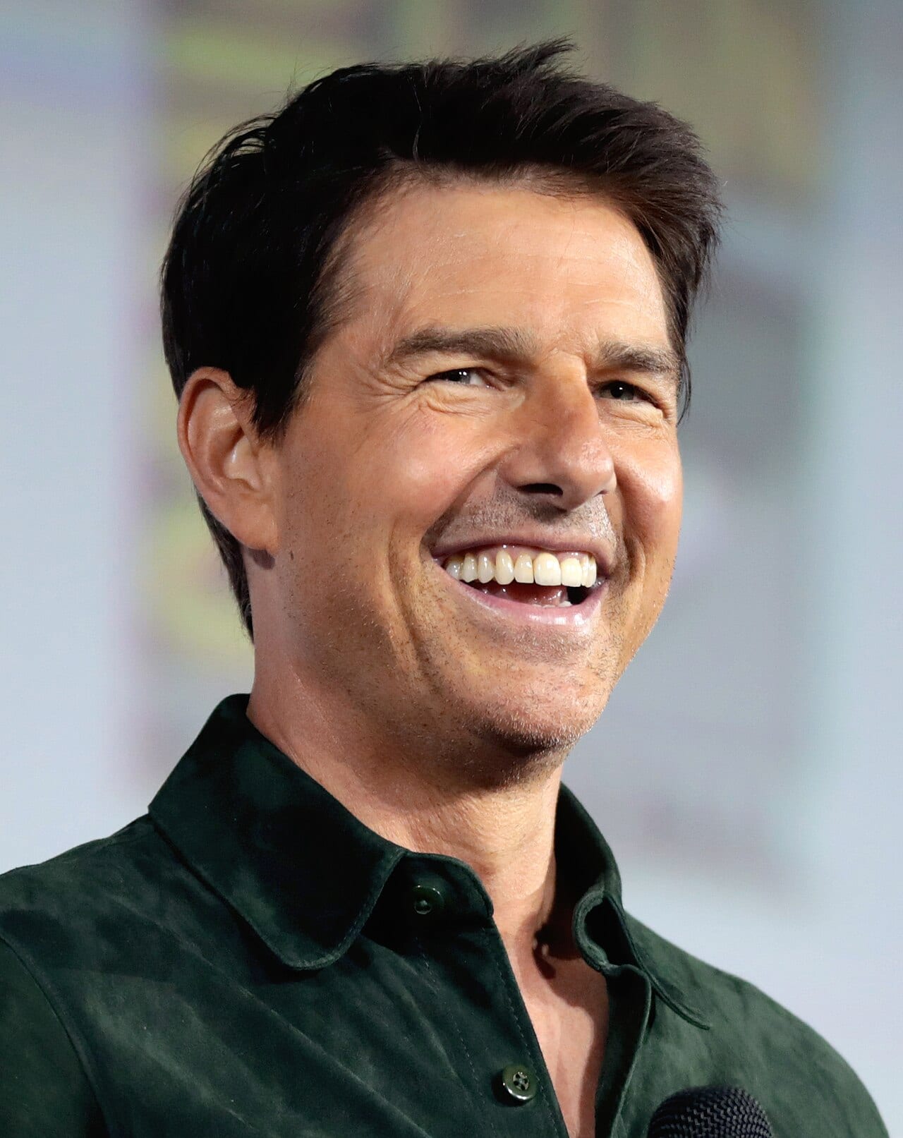 Tom Cruise sonríe de perfil con camisa verde oscura de cuello abotonado, frente a un fondo desenfocado en tonos claros.