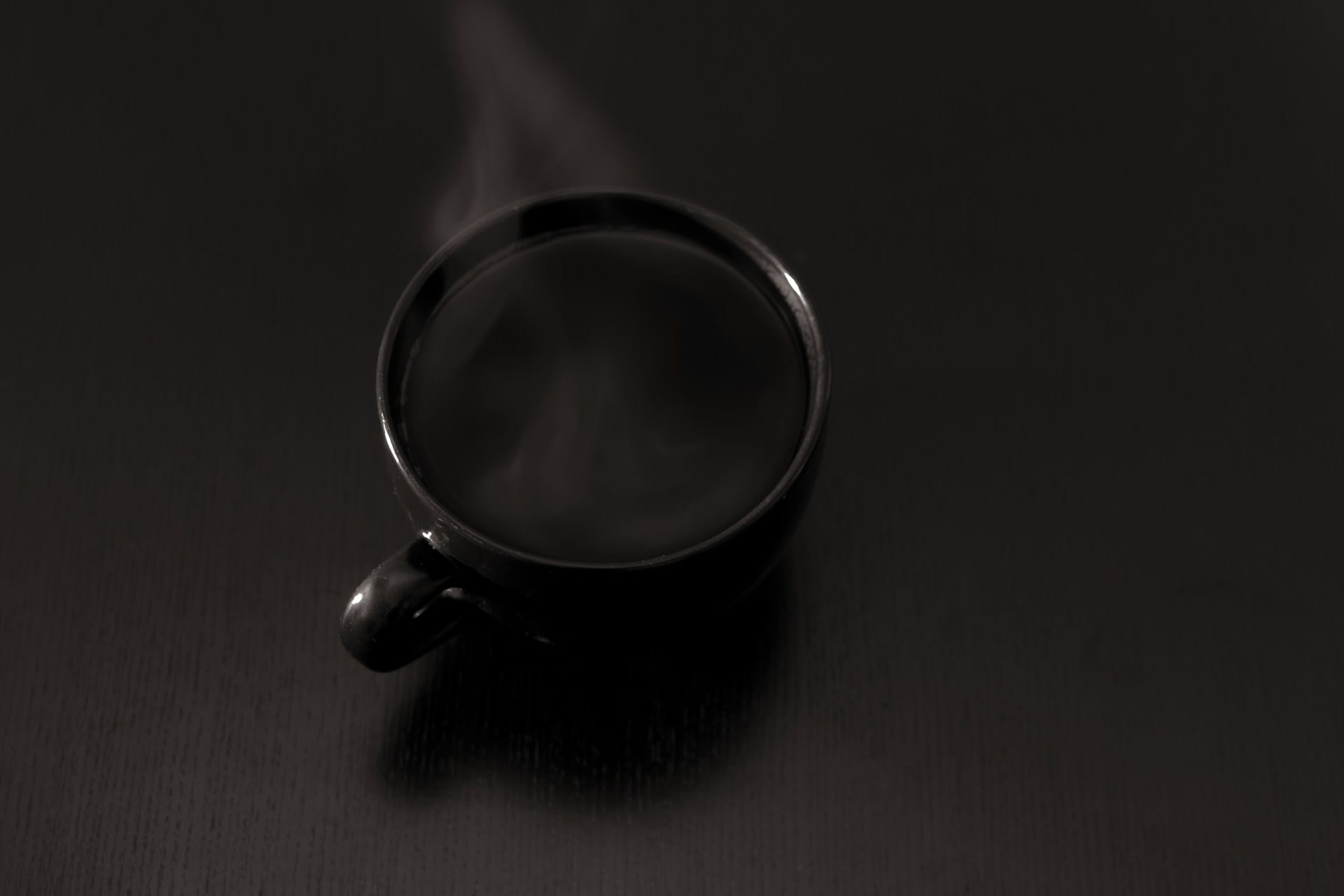 Taza negra con café humeante vista desde arriba, sobre una mesa oscura de madera, con un hilo de vapor que sube hacia el borde superior.