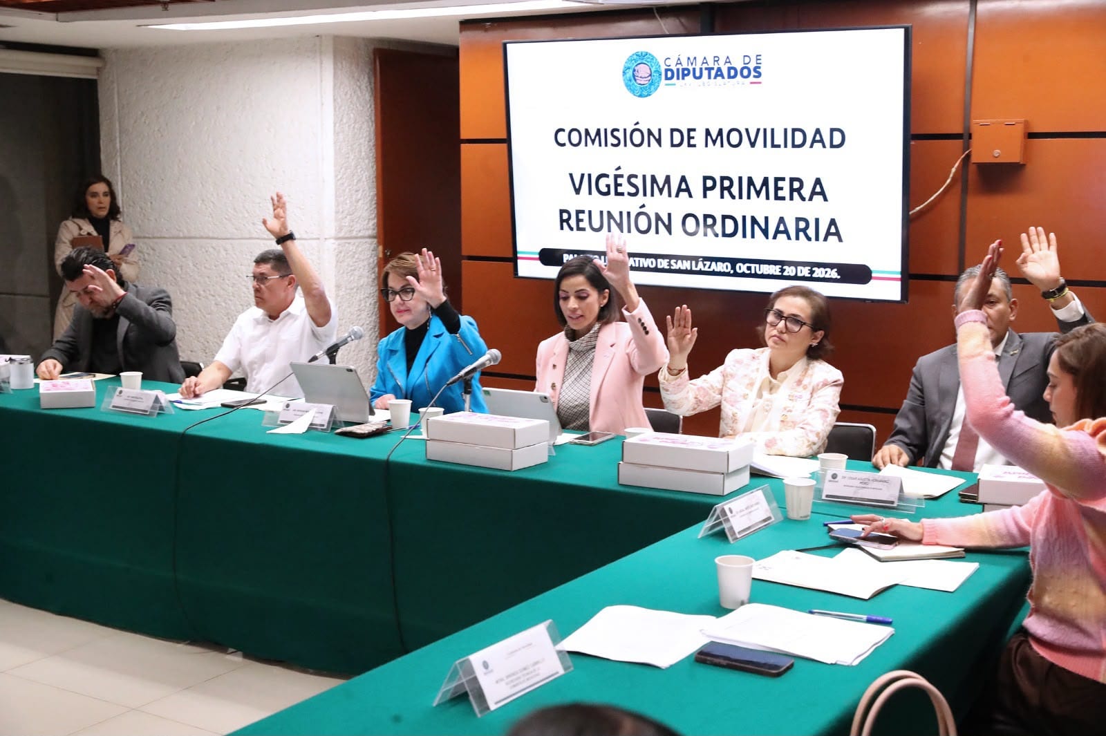 Integrantes de la Comisión de Movilidad levantan la mano para votar en una mesa con mantel verde, bajo una pantalla que anuncia su vigésima primera reunión ordinaria del 20 de octubre de 2026.