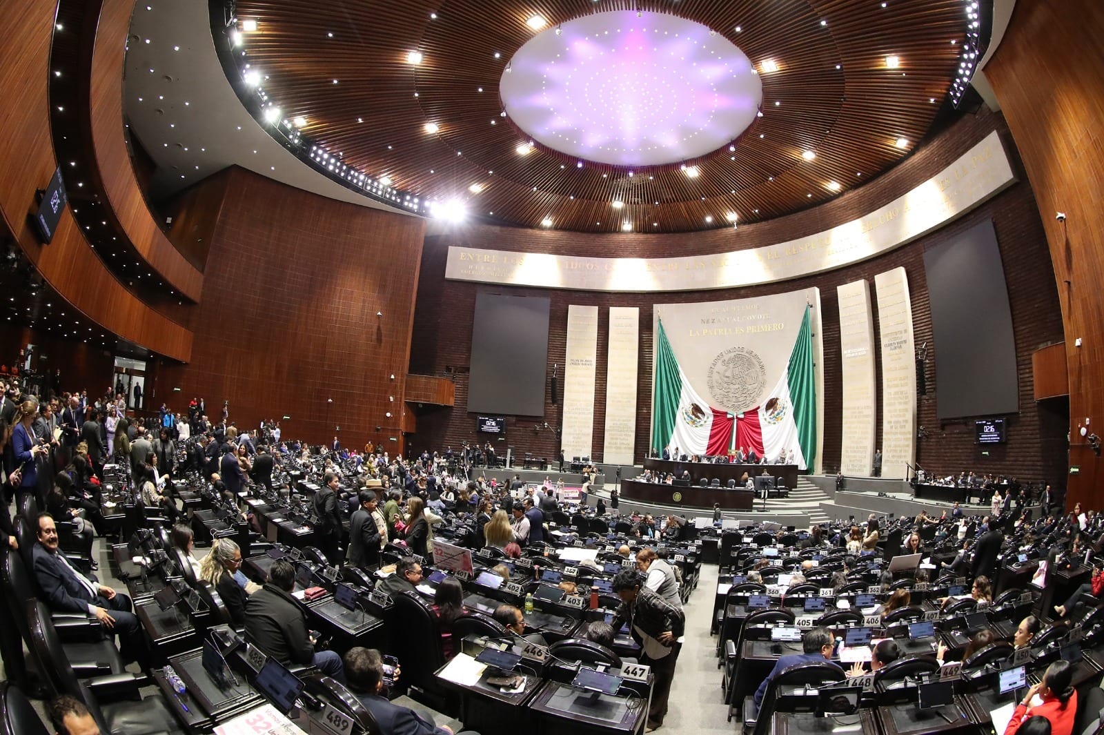 Pleno de la Cámara de Diputados visto desde un costado, con legisladores de pie entre las curules y la bandera nacional detrás de la mesa directiva.