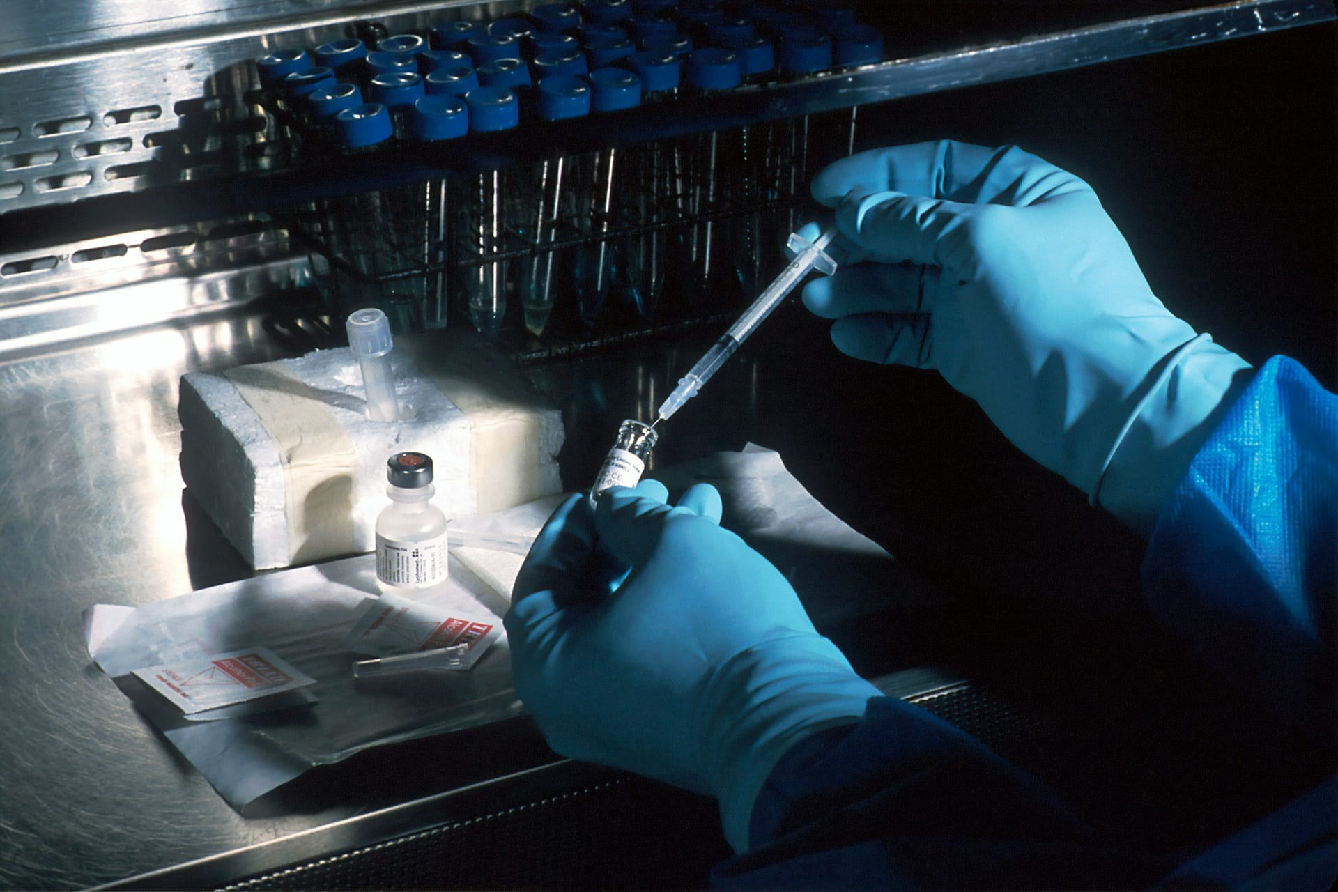 Manos con guantes de látex sostienen una jeringa y un frasco pequeño con líquido mientras extraen el contenido, bajo una campana de laboratorio.