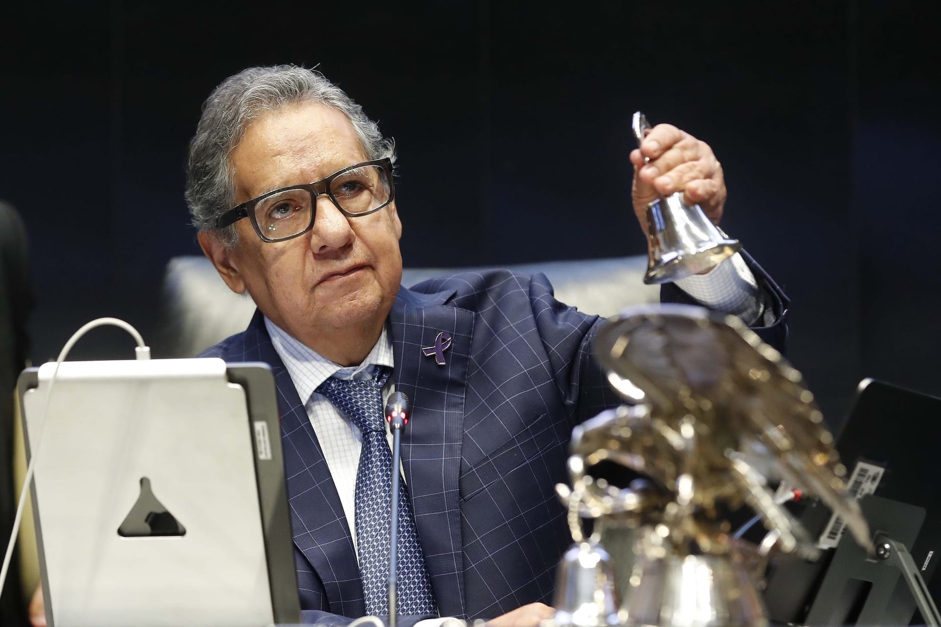 Un hombre de pelo canoso y lentes levanta una campana plateada en un escenario oscuro, junto a un águila metálica y un micrófono.