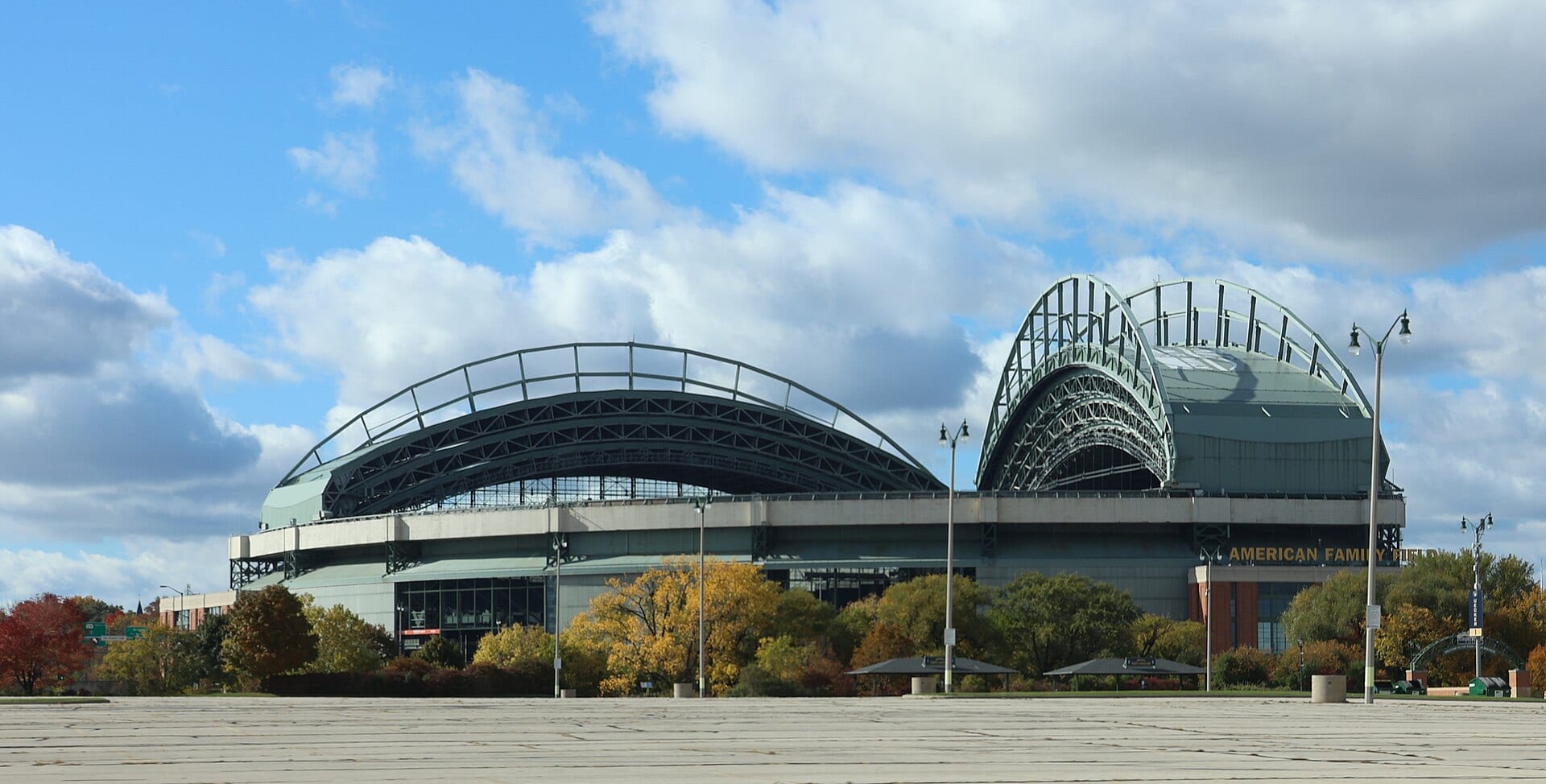 Fachada del American Family Field con su techo retráctil abierto en dos arcos, árboles de otoño y un estacionamiento vacío en primer plano.