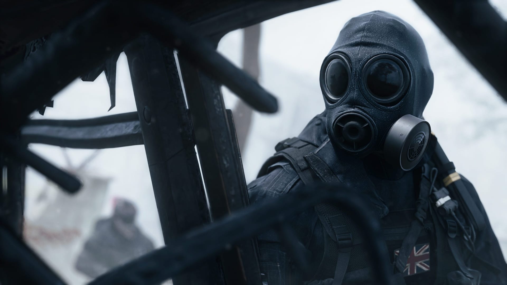A masked operator in a black gas mask with a British flag patch on his vest looks at the camera through twisted metal, with white fog behind him.