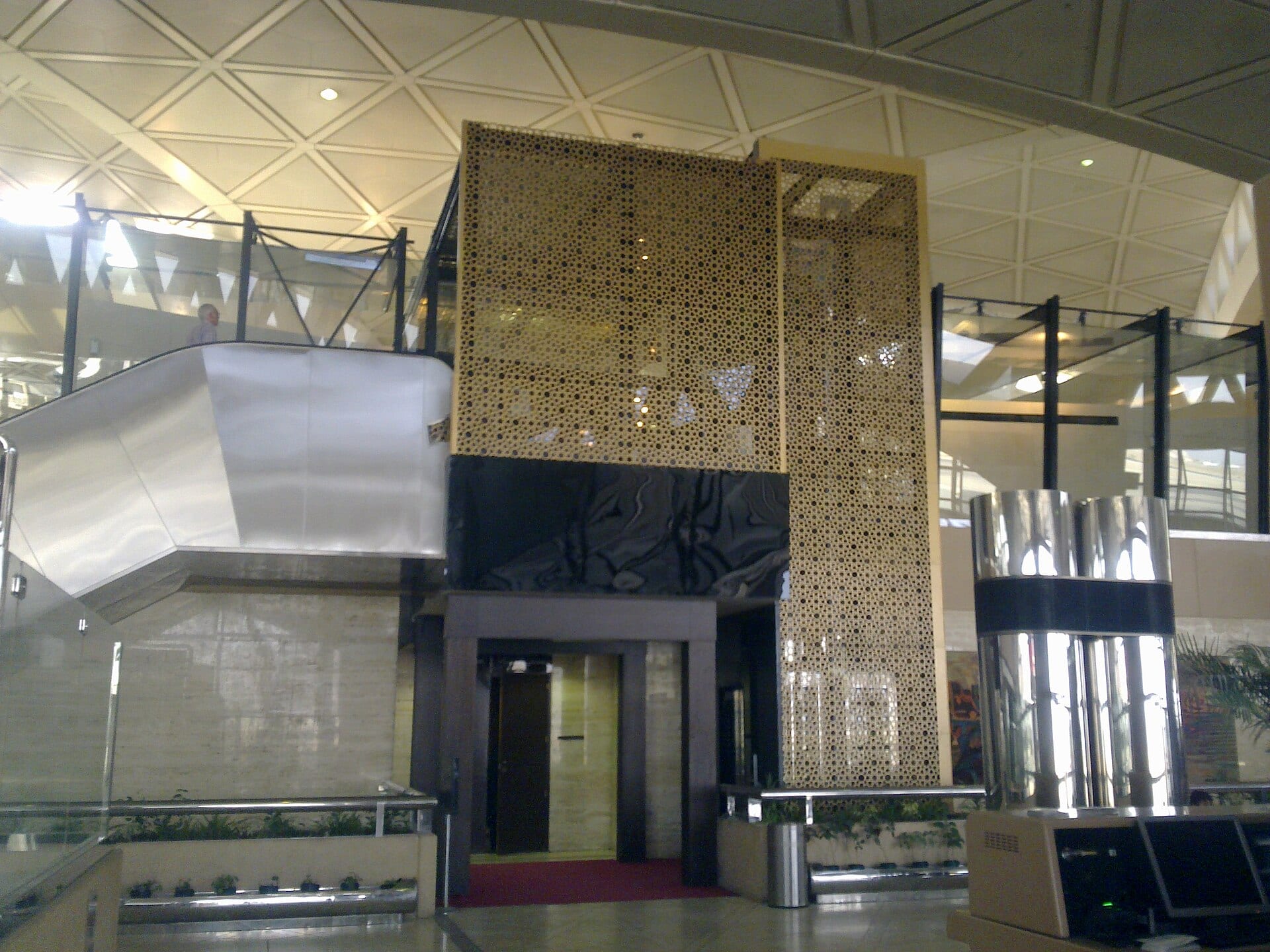 Interior hall of an airport terminal with a white triangular ceiling, an escalator and a gold perforated wall.