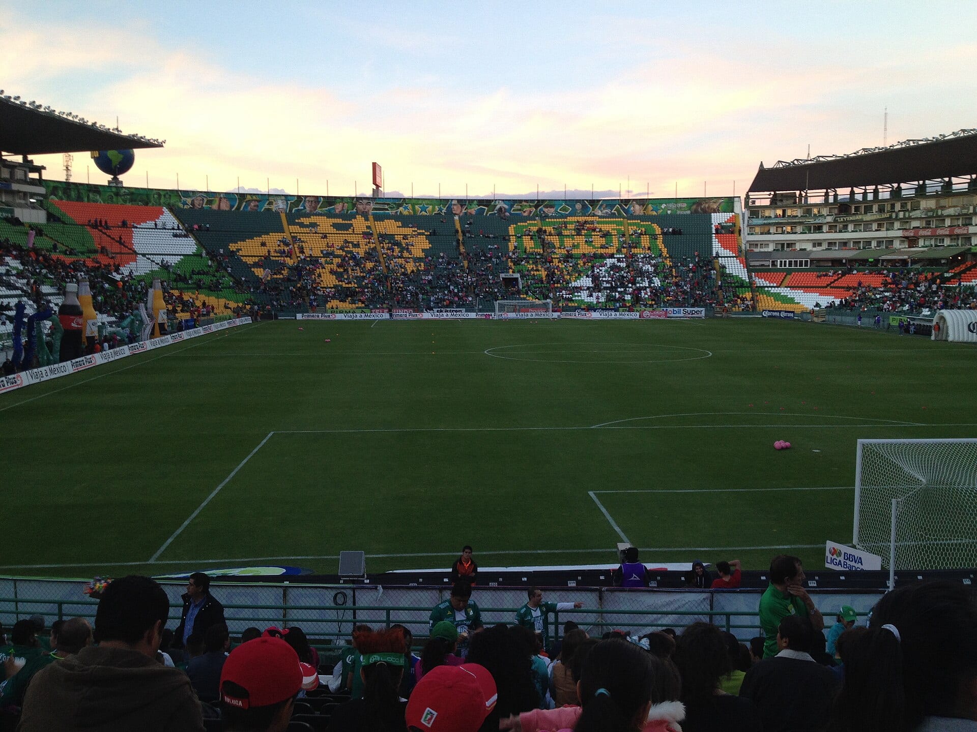 Vista de la cancha del Estadio León desde la tribuna baja, con las gradas pintadas en verde, blanco y naranja y público en las plateas.
