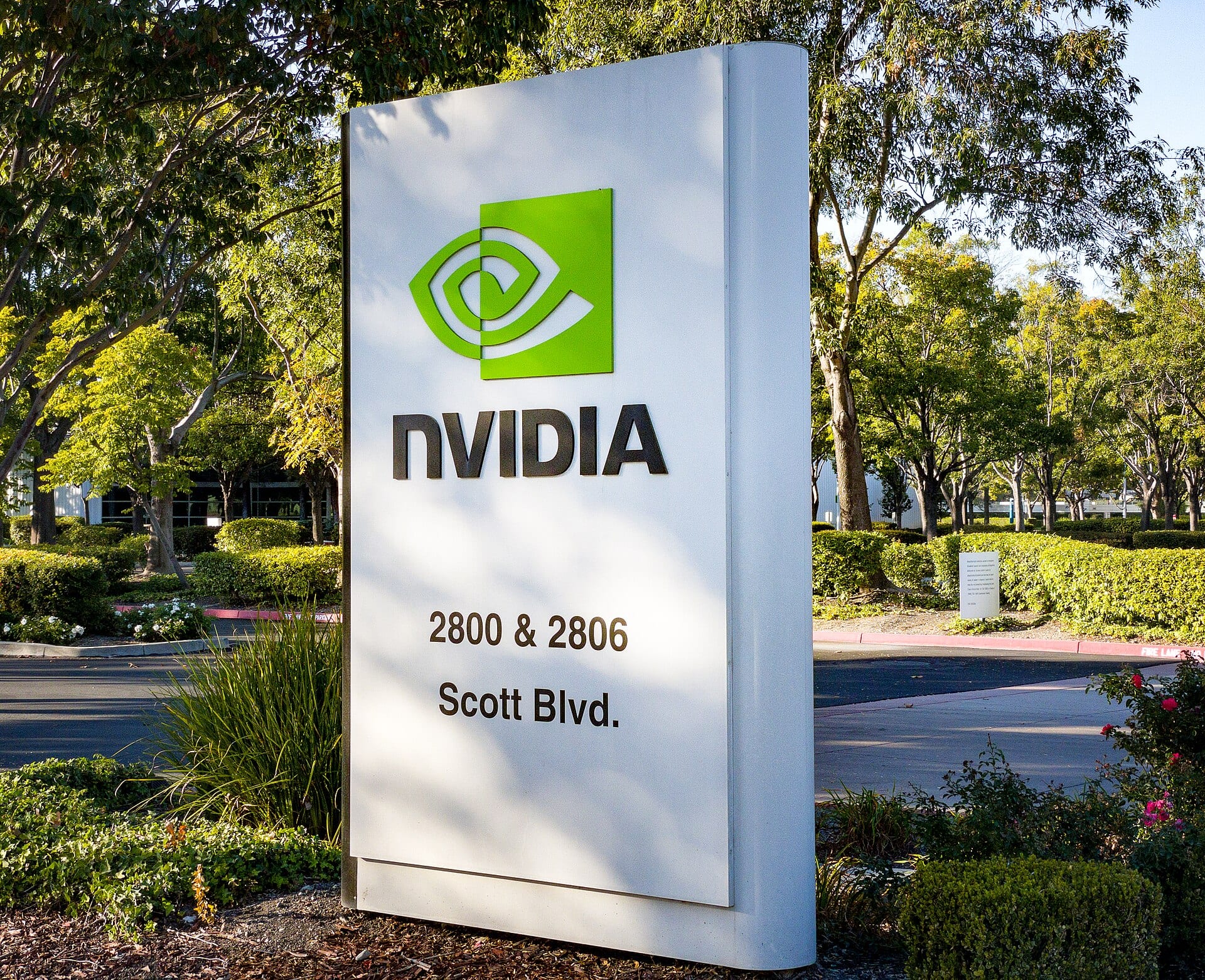 Letrero blanco de Nvidia con su logotipo verde y la dirección 2800 y 2806 Scott Blvd., rodeado de árboles y arbustos.
