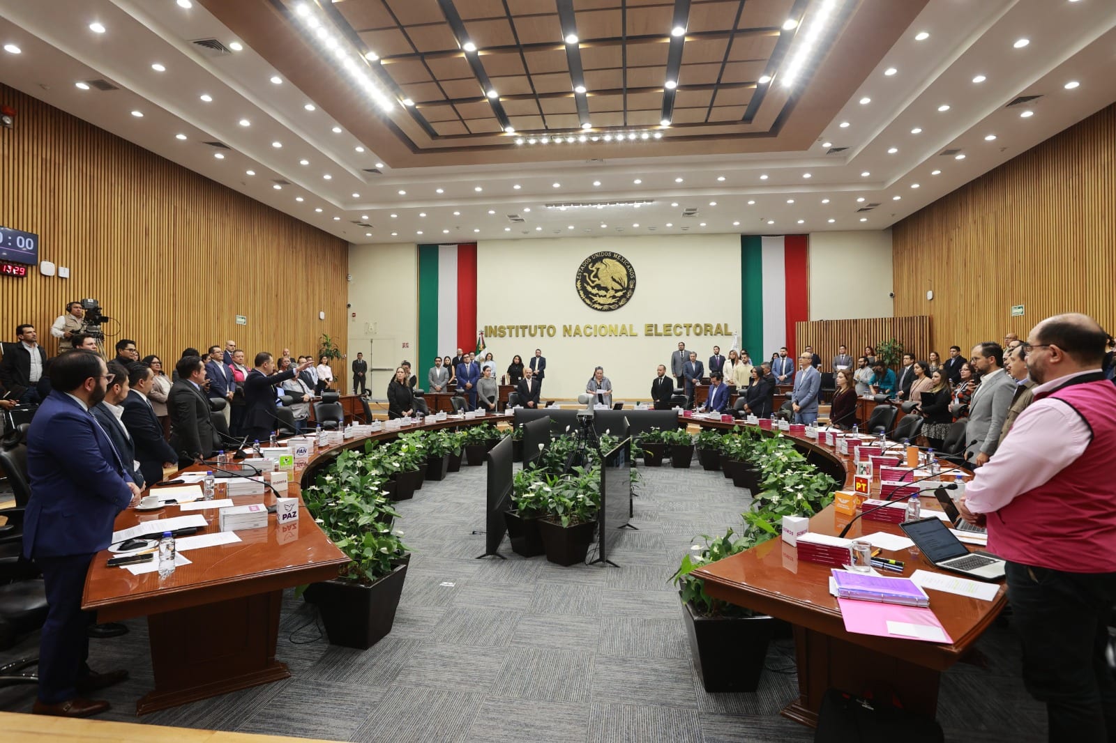 Consejeros y representantes de partidos, de pie, rodean una mesa en herradura en la sala del Consejo General del INE, con banderas de México al fondo.