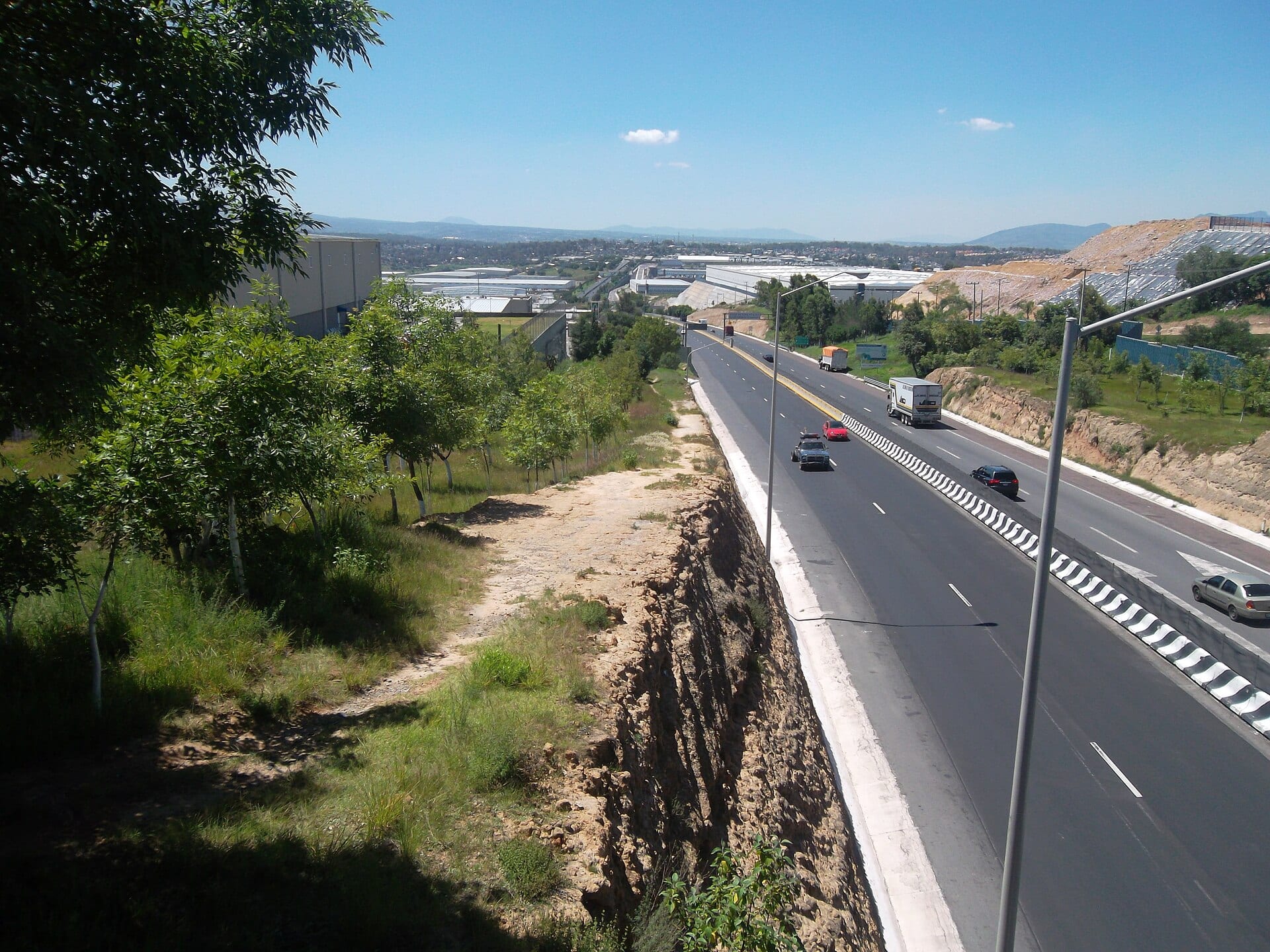 Vista desde lo alto de una carretera de varios carriles con camiones y autos, un talud a la izquierda y naves industriales al fondo.