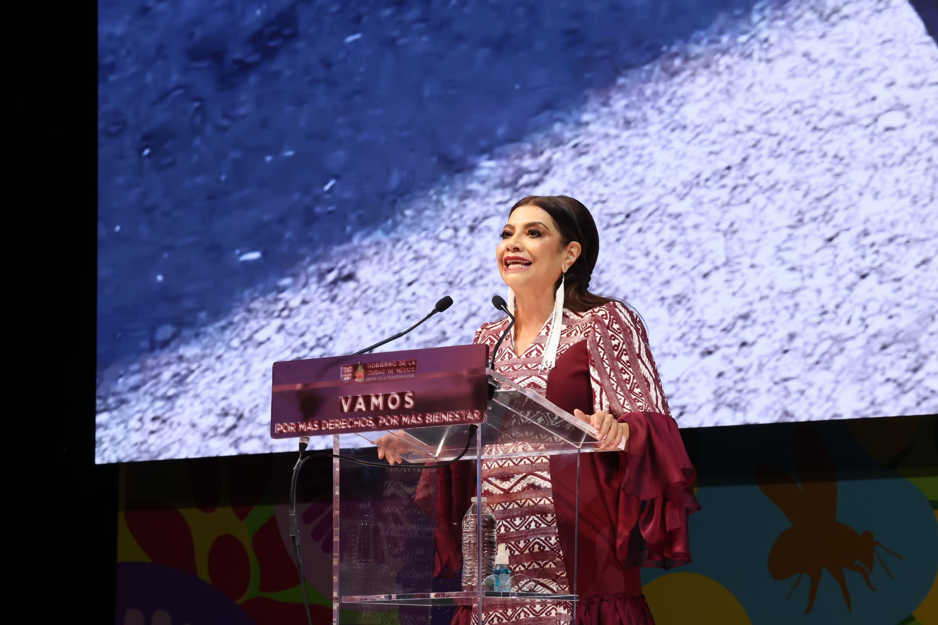 Una mujer de vestido guinda con bordados habla desde un podio de acrílico frente a una pantalla, en un escenario con fondo de flores de colores.