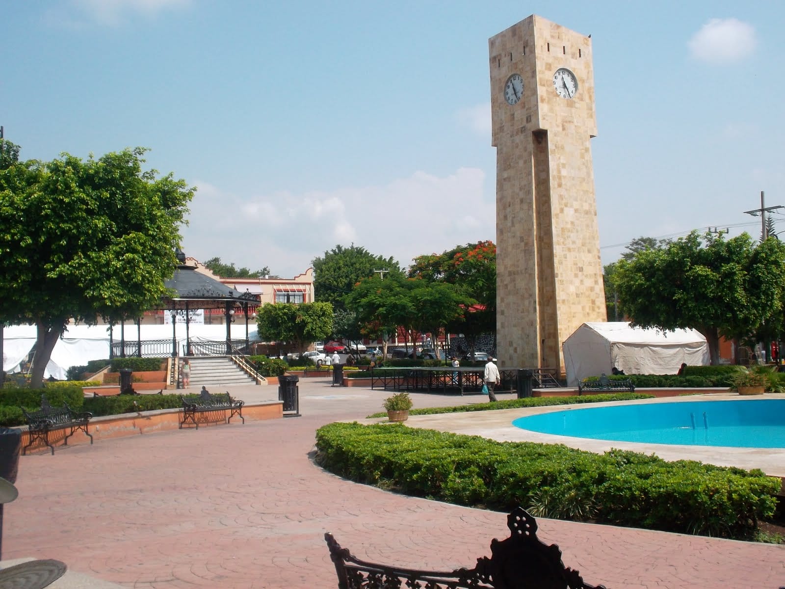 Una torre de reloj de piedra clara se alza junto a un quiosco y una fuente, en una plaza con árboles y bancas de hierro de Jiutepec.