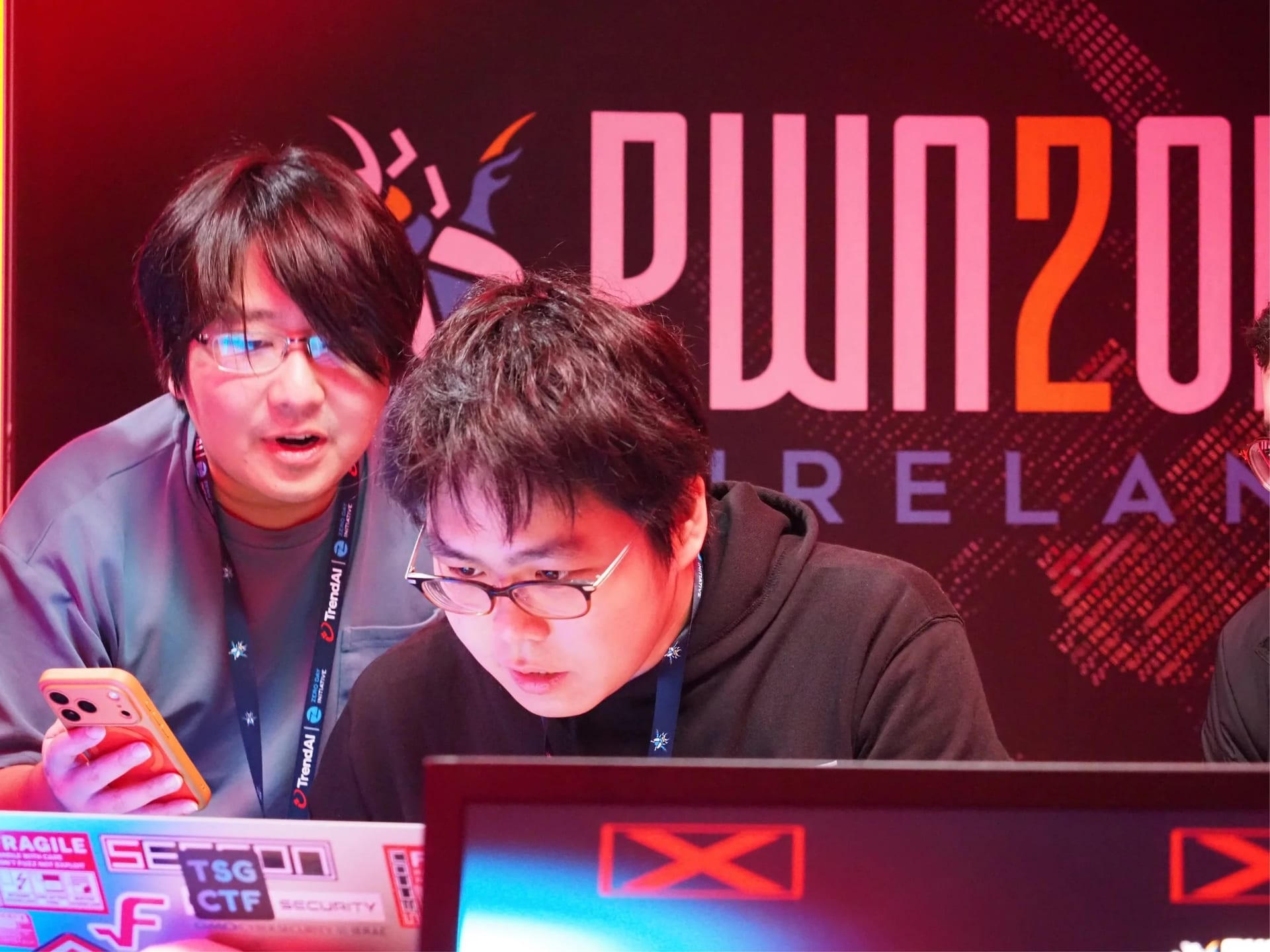 Two contestants look at a laptop screen in front of the Pwn2Own Ireland sign.