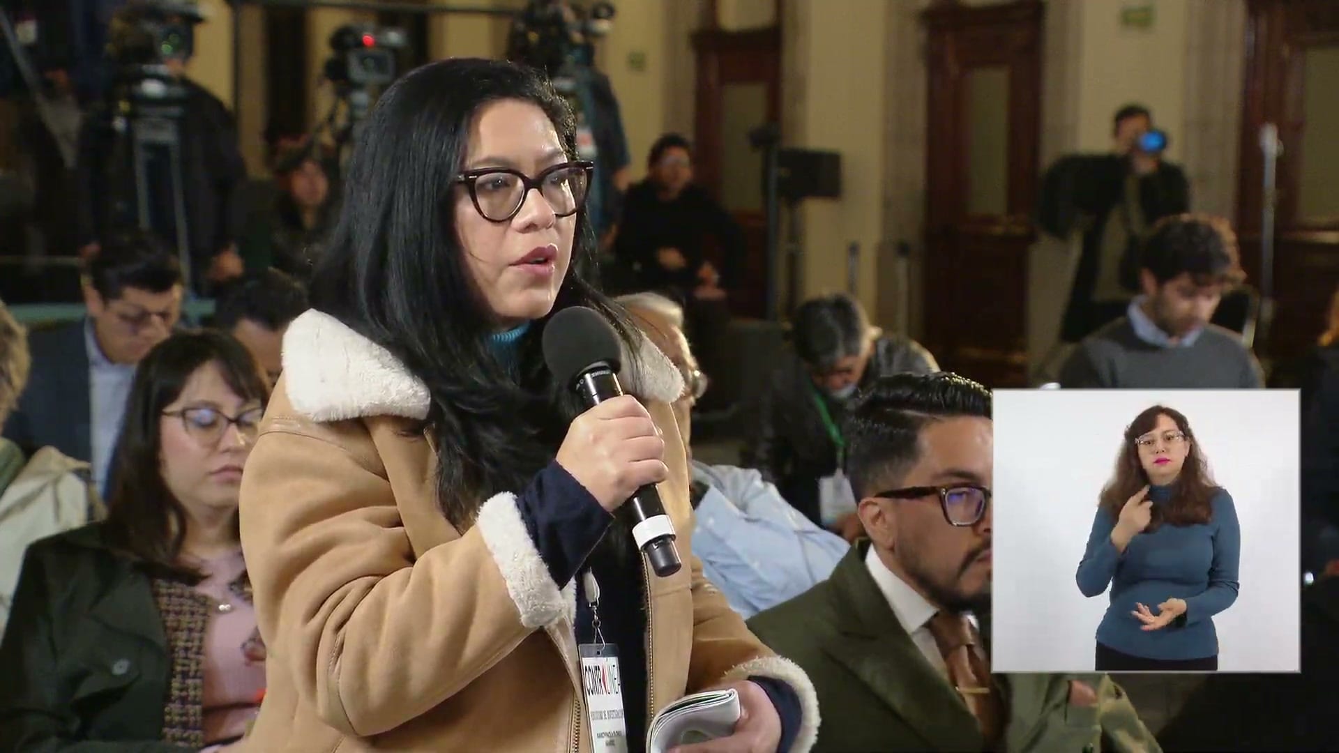 Una periodista de pie sostiene un micrófono y hace una pregunta en la conferencia matutina; a la derecha, una intérprete de lengua de señas.