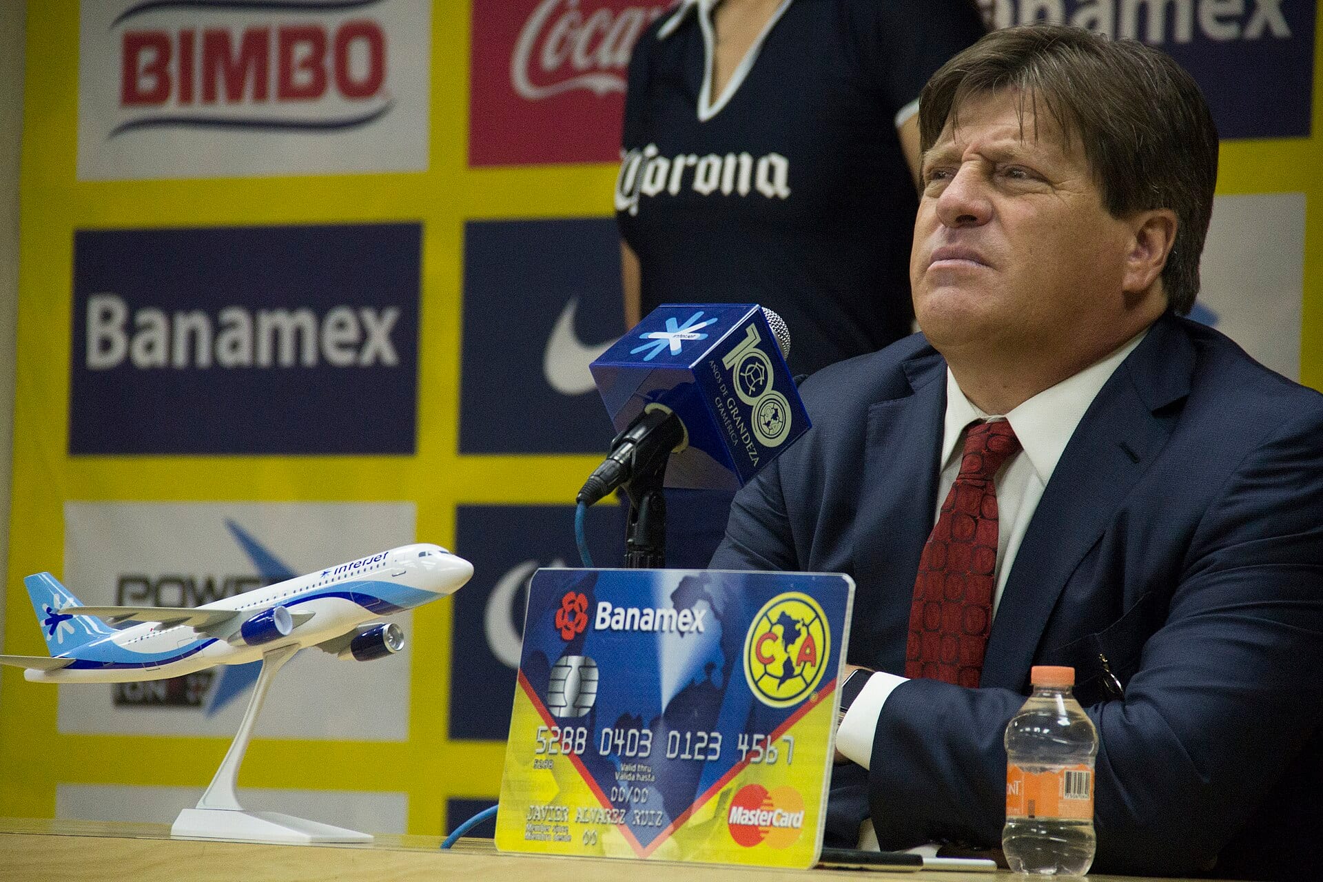 Miguel Herrera, de traje azul y corbata roja, escucha sentado en una conferencia de prensa con un micrófono y patrocinadores del América detrás.