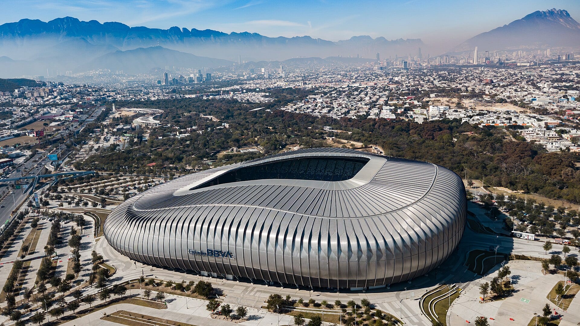 Vista aérea del estadio de Monterrey, de fachada metálica, con la ciudad y las montañas al fondo.
