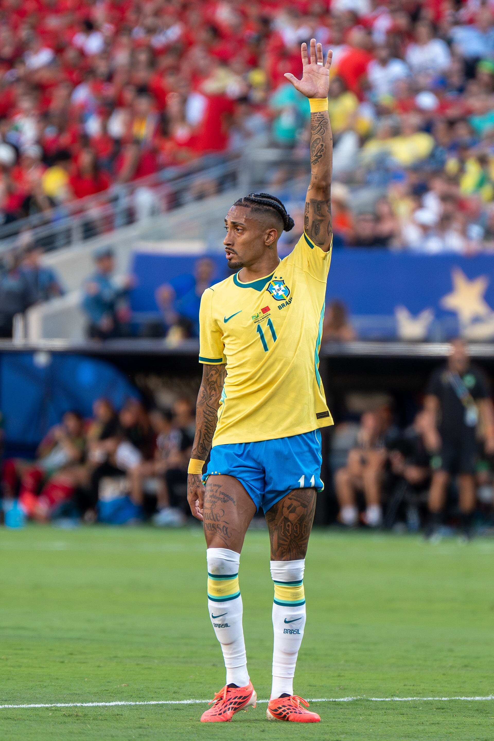 Raphinha, con la camiseta amarilla de Brasil y el número 11, levanta la mano derecha en el campo con las gradas al fondo.