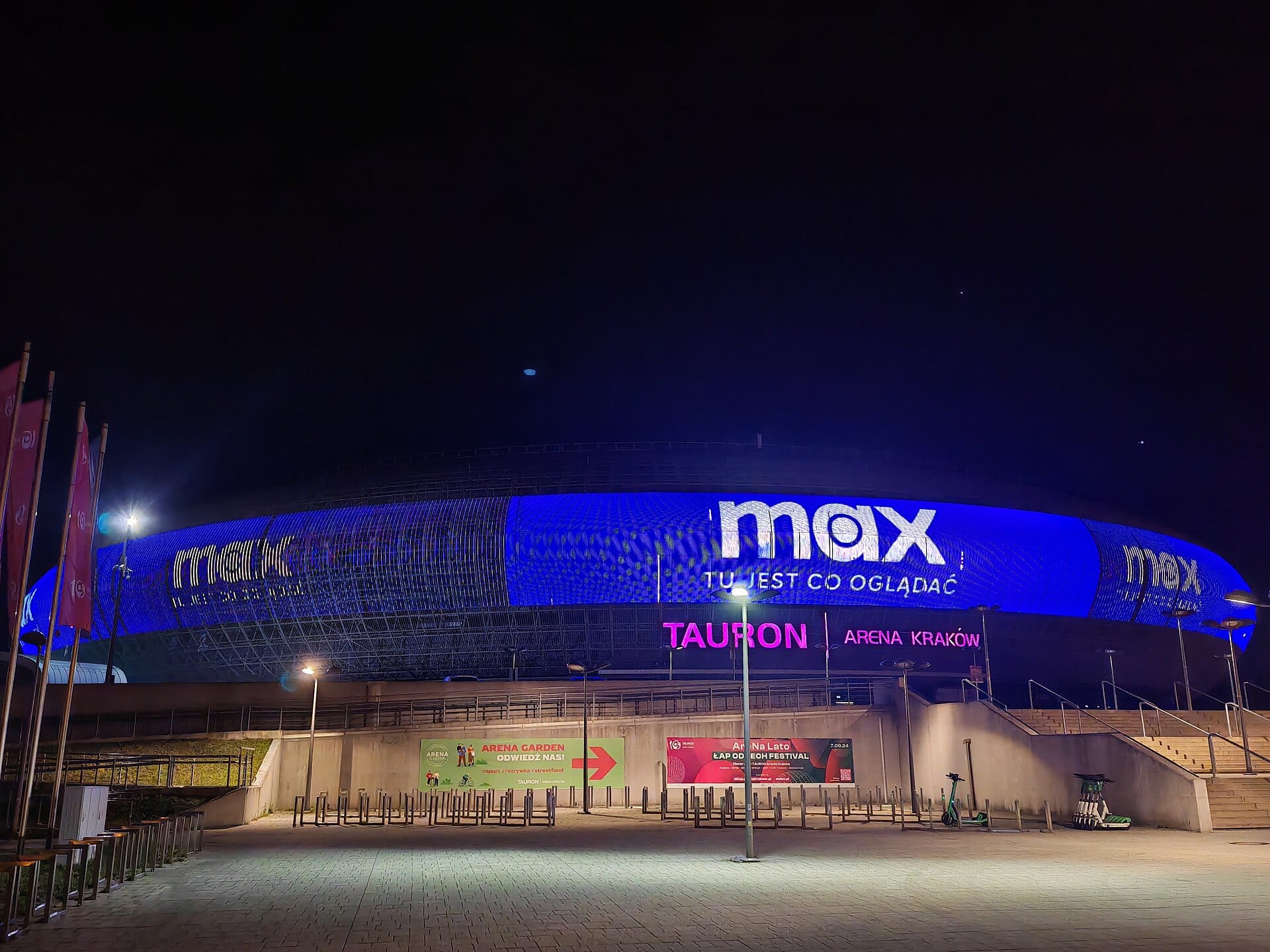 Fachada iluminada en azul de un recinto deportivo de noche, con el logotipo de la plataforma Max proyectado sobre el anillo exterior del edificio.